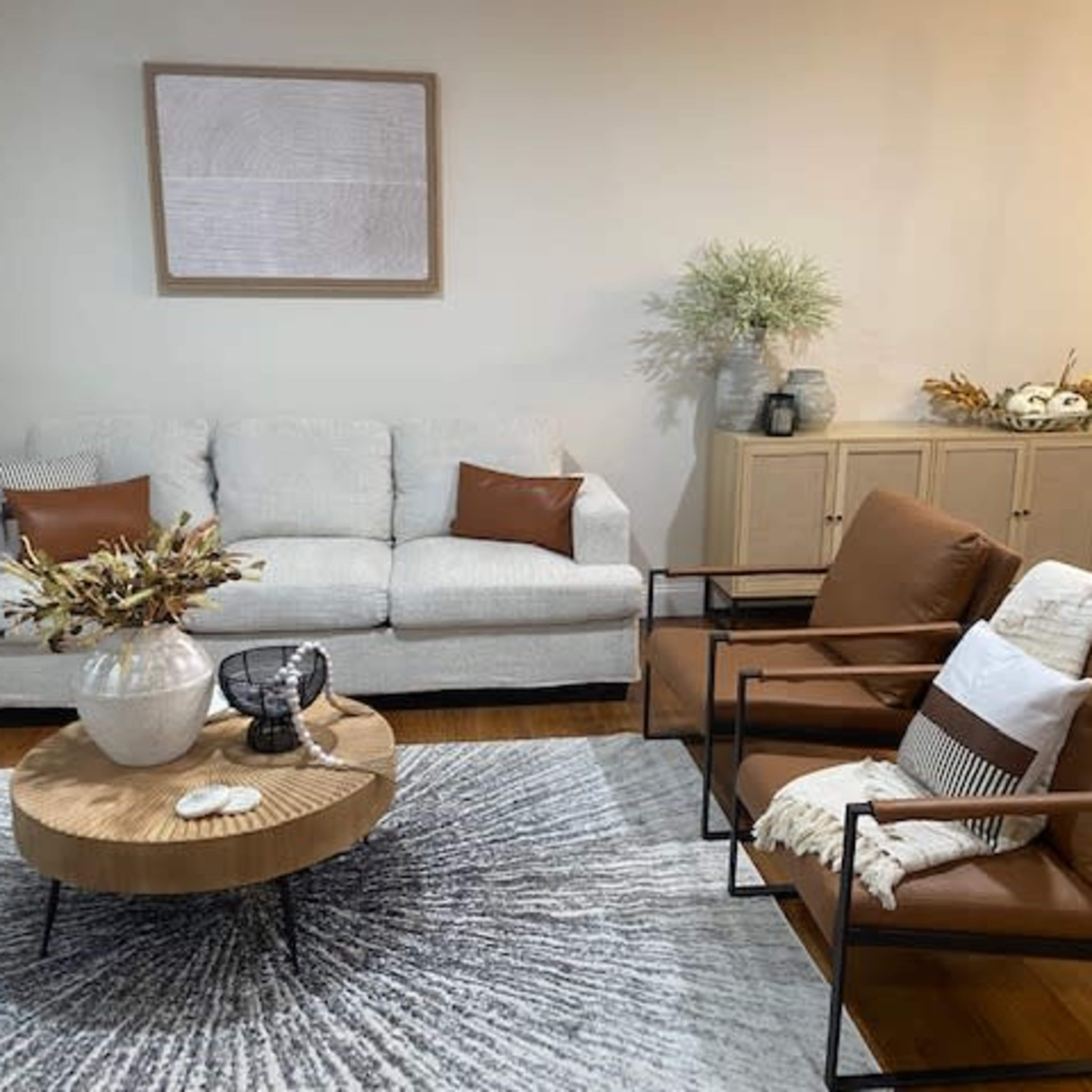 A living room with a light-colored sofa, two brown chairs, a round coffee table, and decorative elements on a large circular rug.