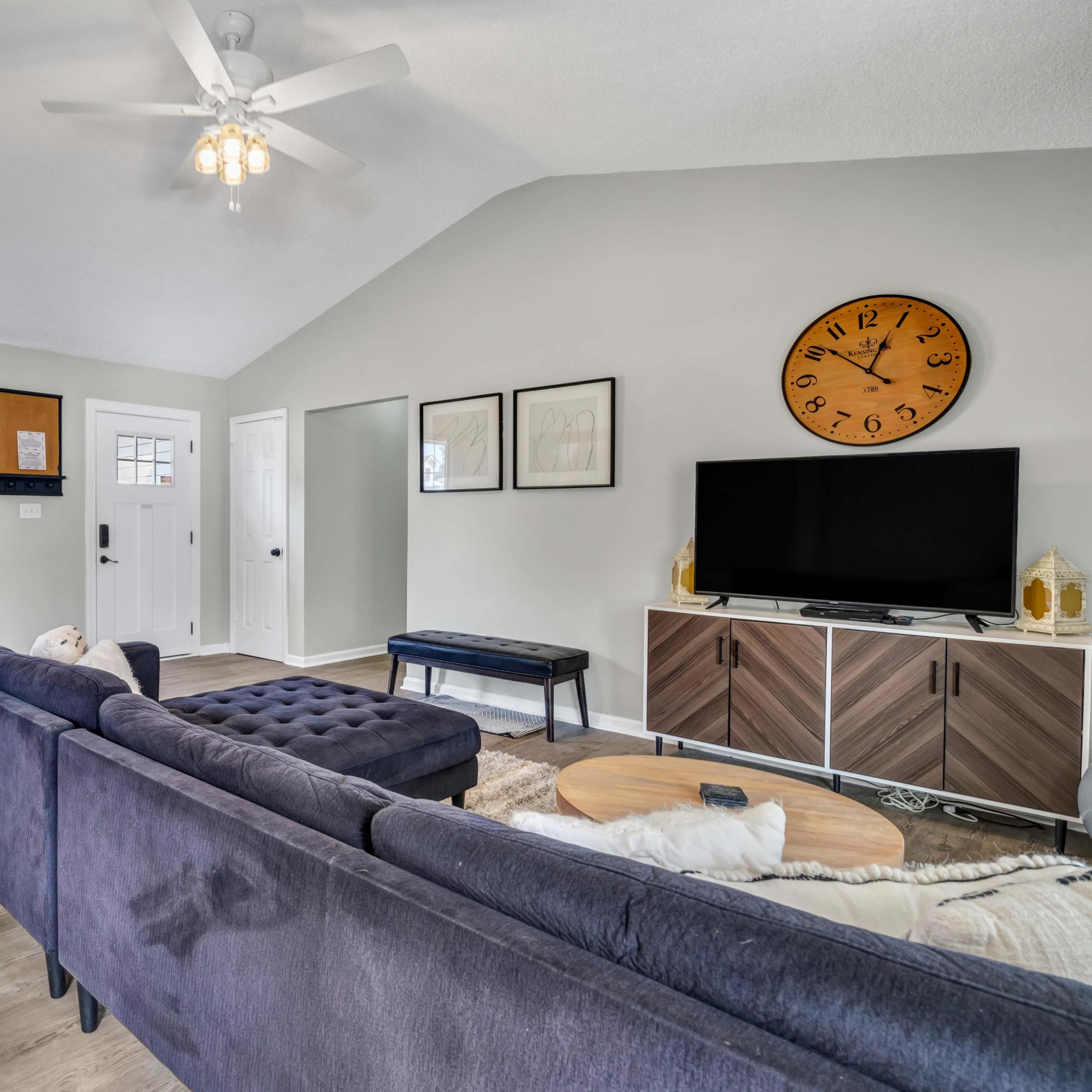 A modern living room features a sectional sofa, a coffee table, and a television on a low cabinet, illuminated by natural light from a large window.