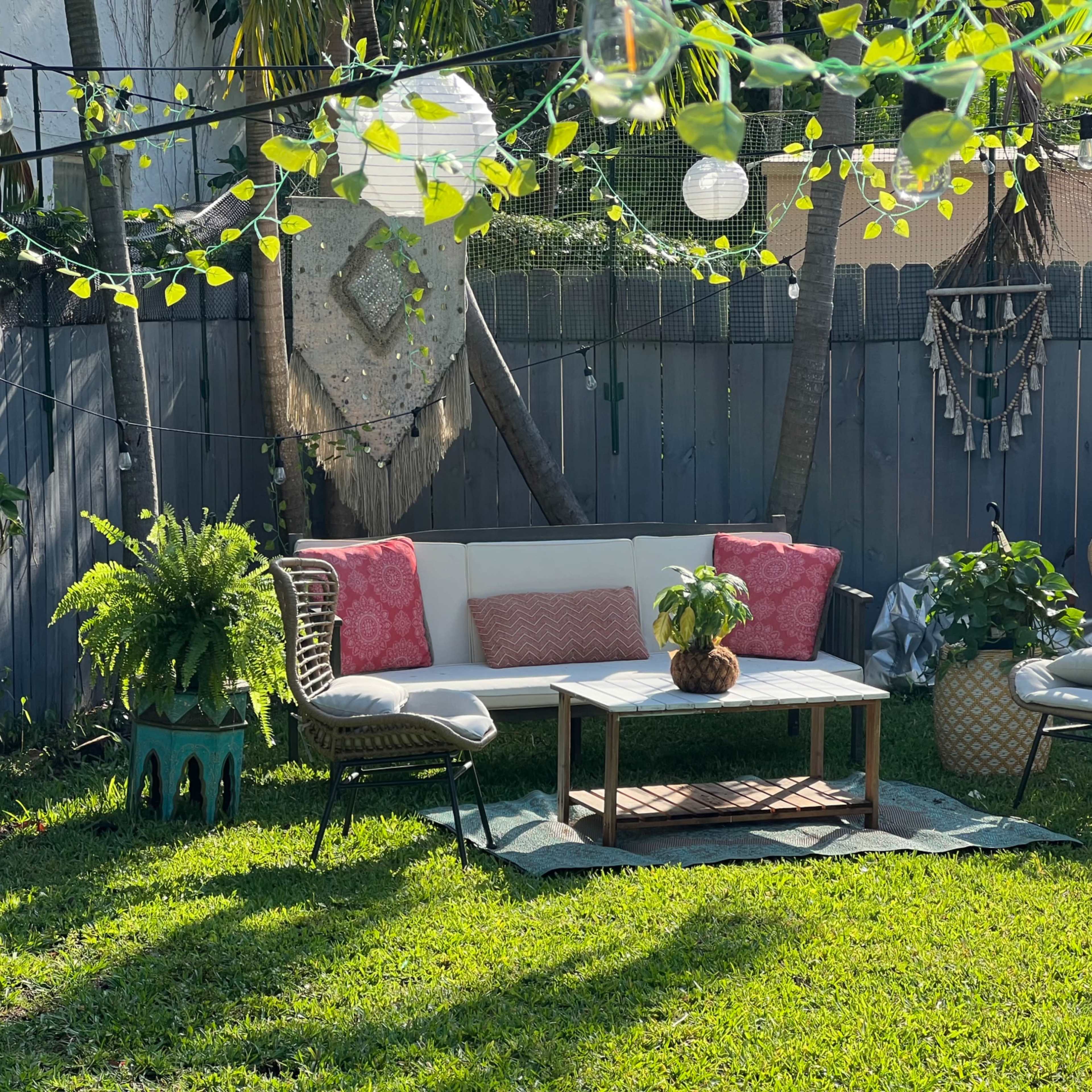 A cozy backyard seating area features a white sectional sofa, decorative pillows, a coffee table, and soft lighting among lush greenery.