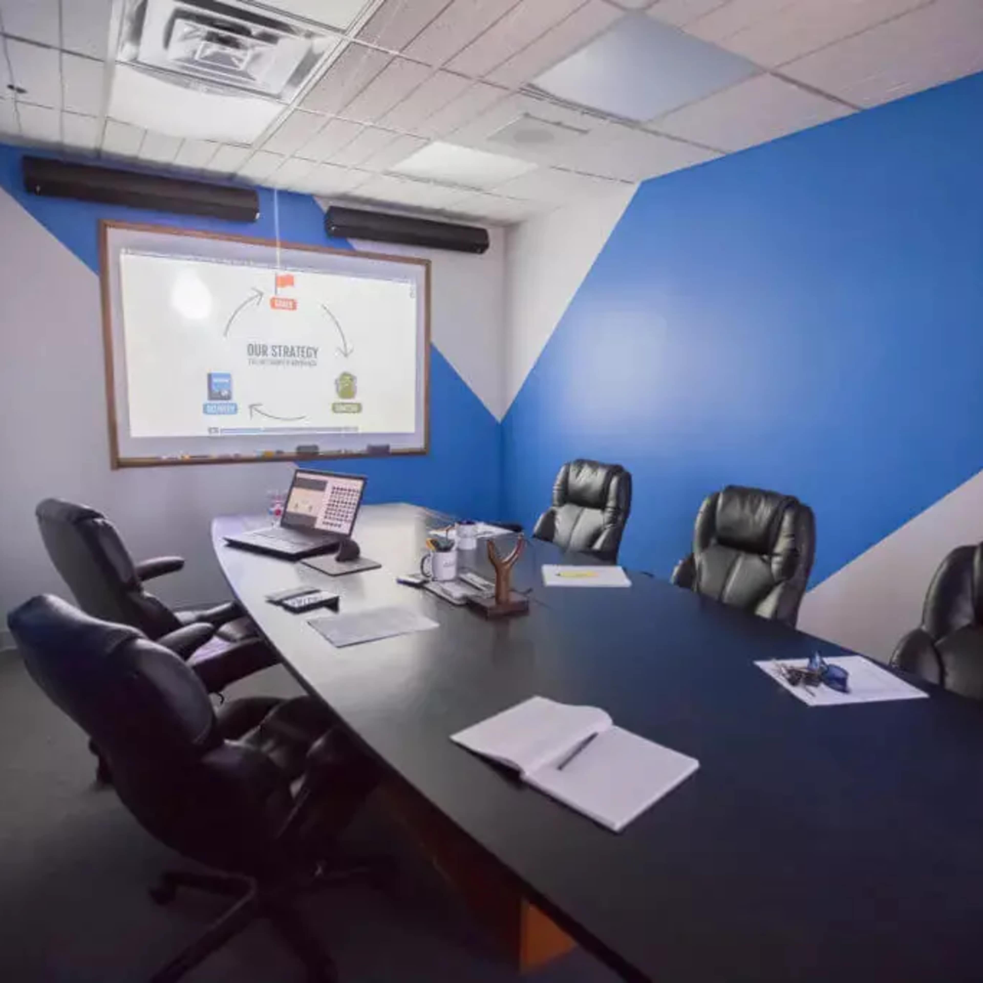 A conference room with a large table, several black office chairs, a laptop, documents, and a projector screen displaying a flowchart against a blue and gray wall.