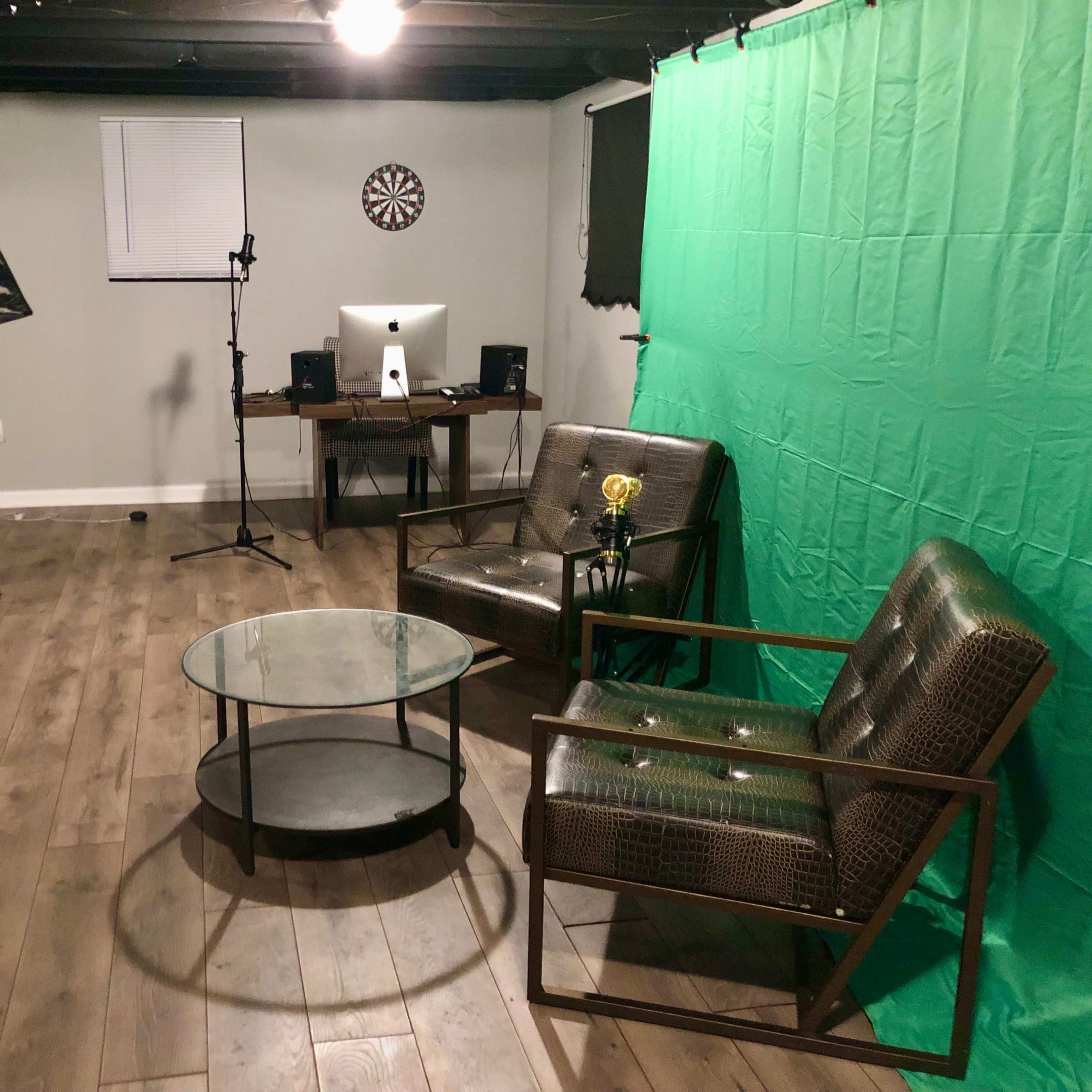 A basement studio setup with a green screen, two chairs, a glass coffee table, and equipment for recording or broadcasting.