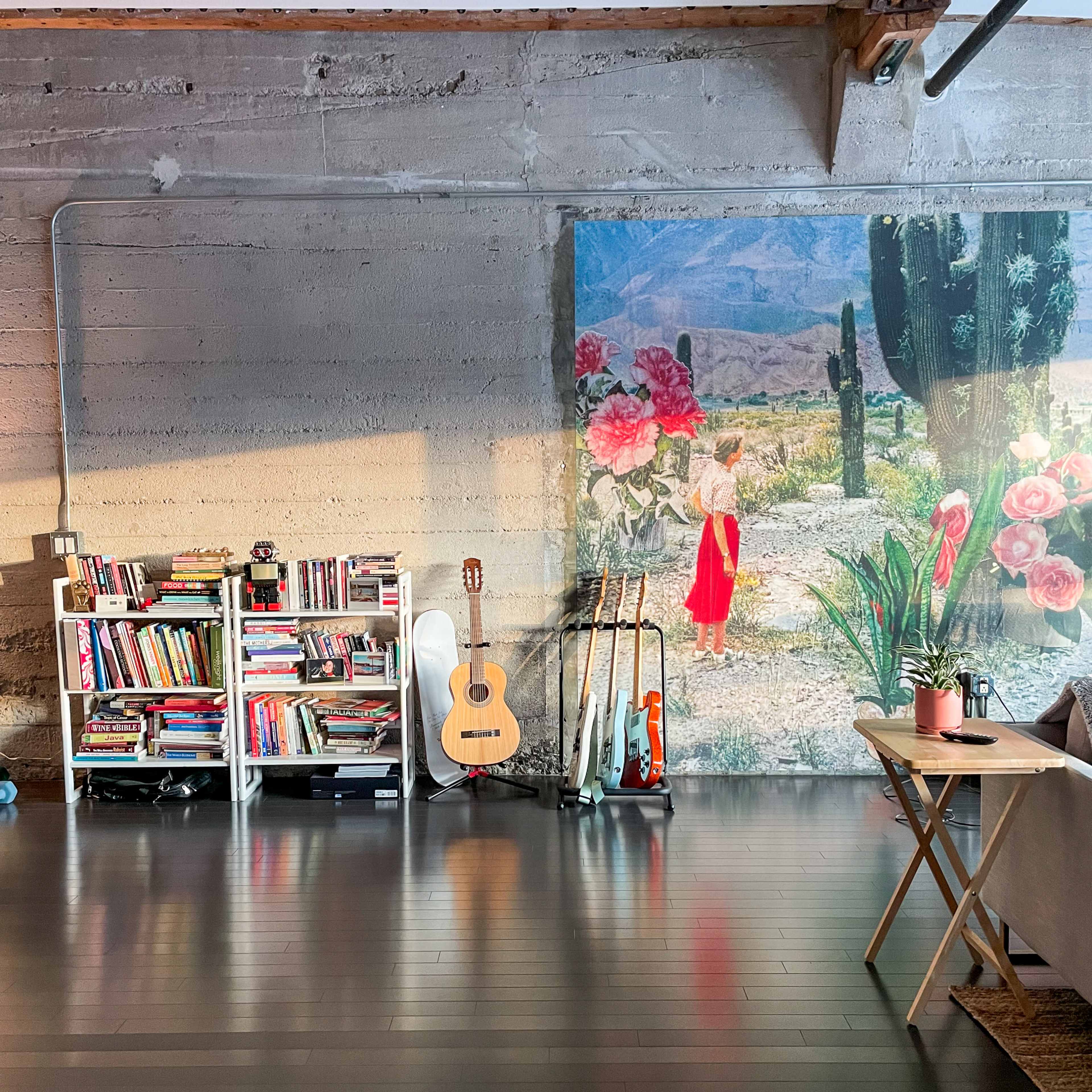 A cozy room features a bookshelf filled with books, an acoustic guitar, several surfboards, and a large painting of a desert scene with a person amidst cacti and flowers.