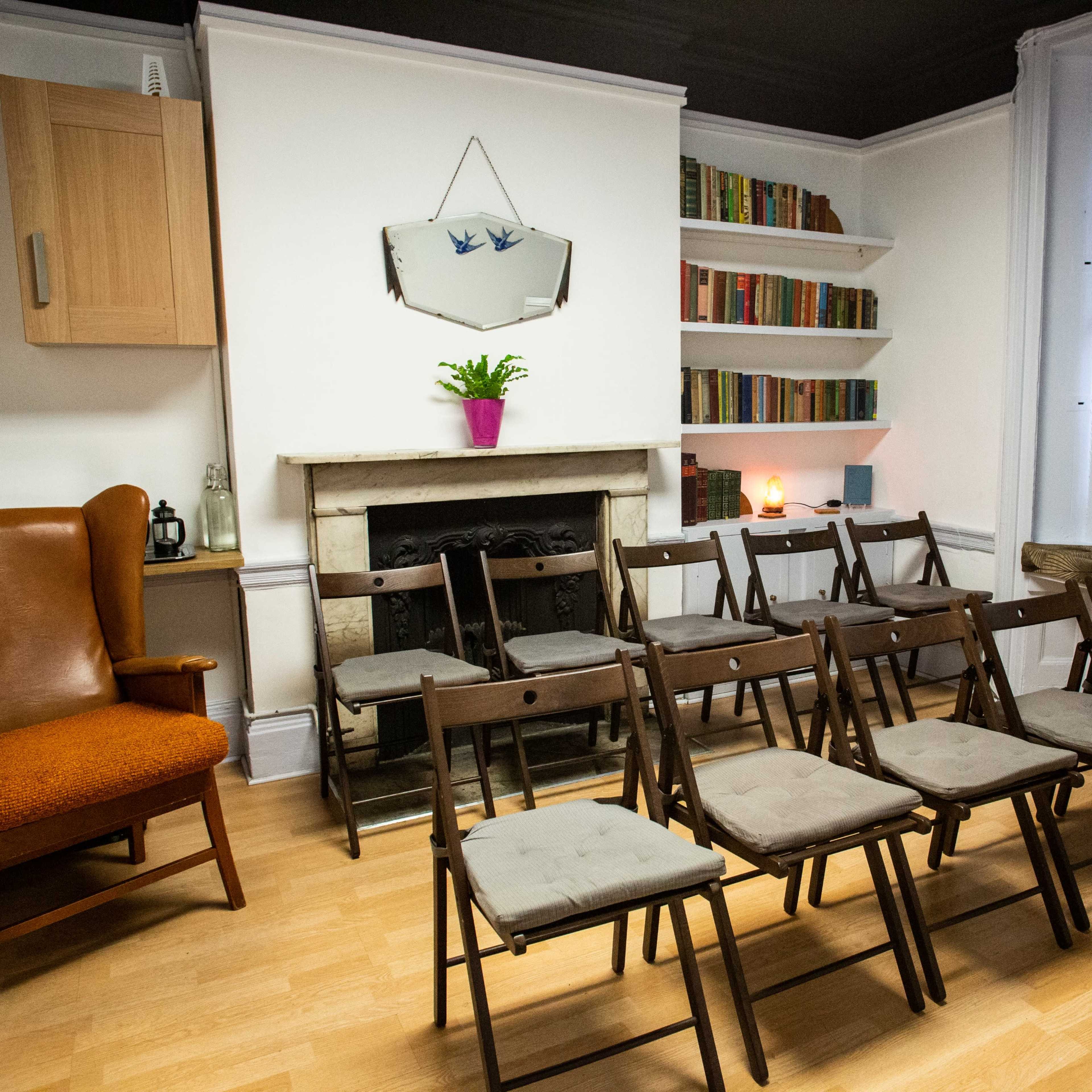 A small room features a row of folding chairs facing a mantel with a mirror, a bookshelf filled with books, and a large armchair in the corner.