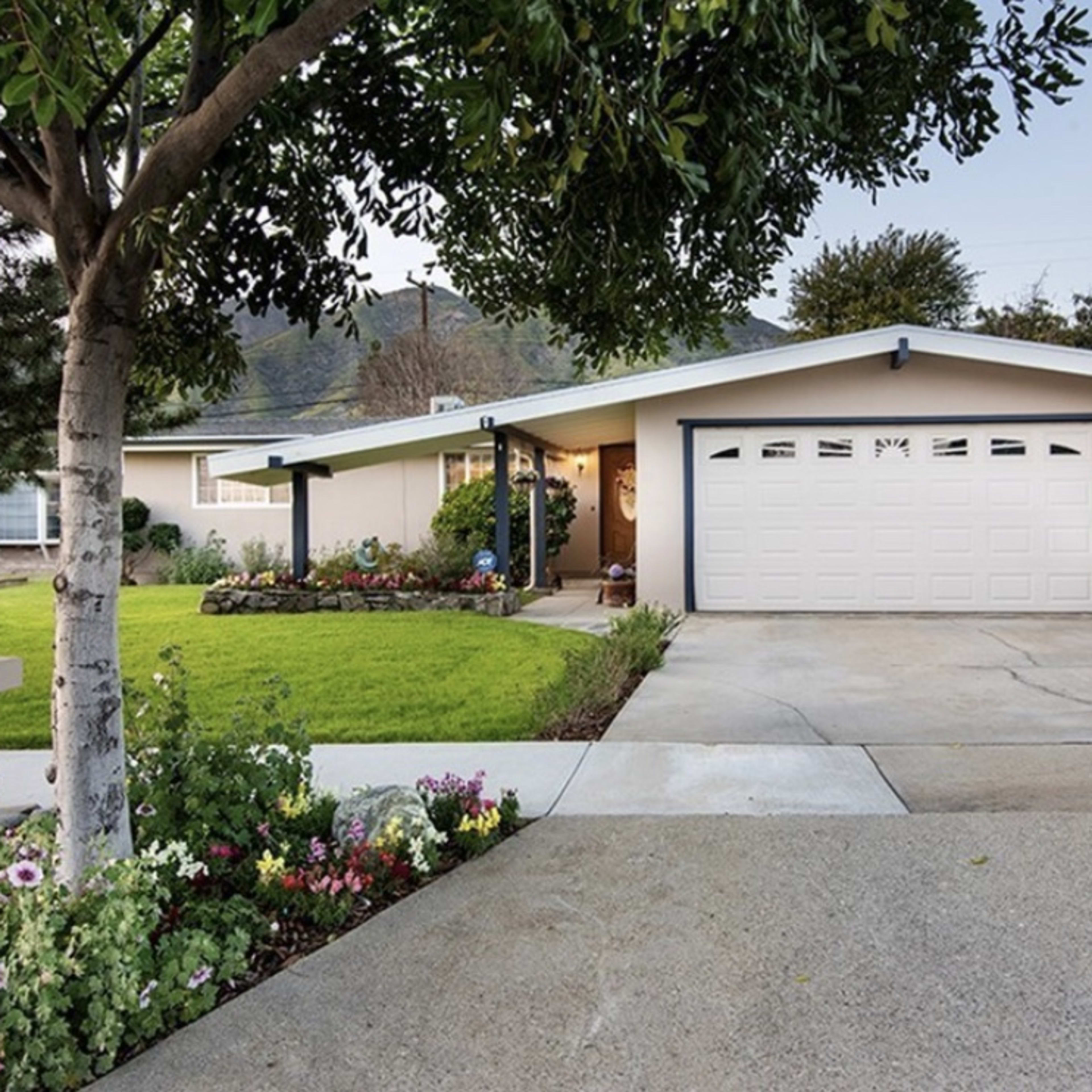 A single-story house with a two-car garage is set on a manicured lawn, surrounded by flower beds and a tree.