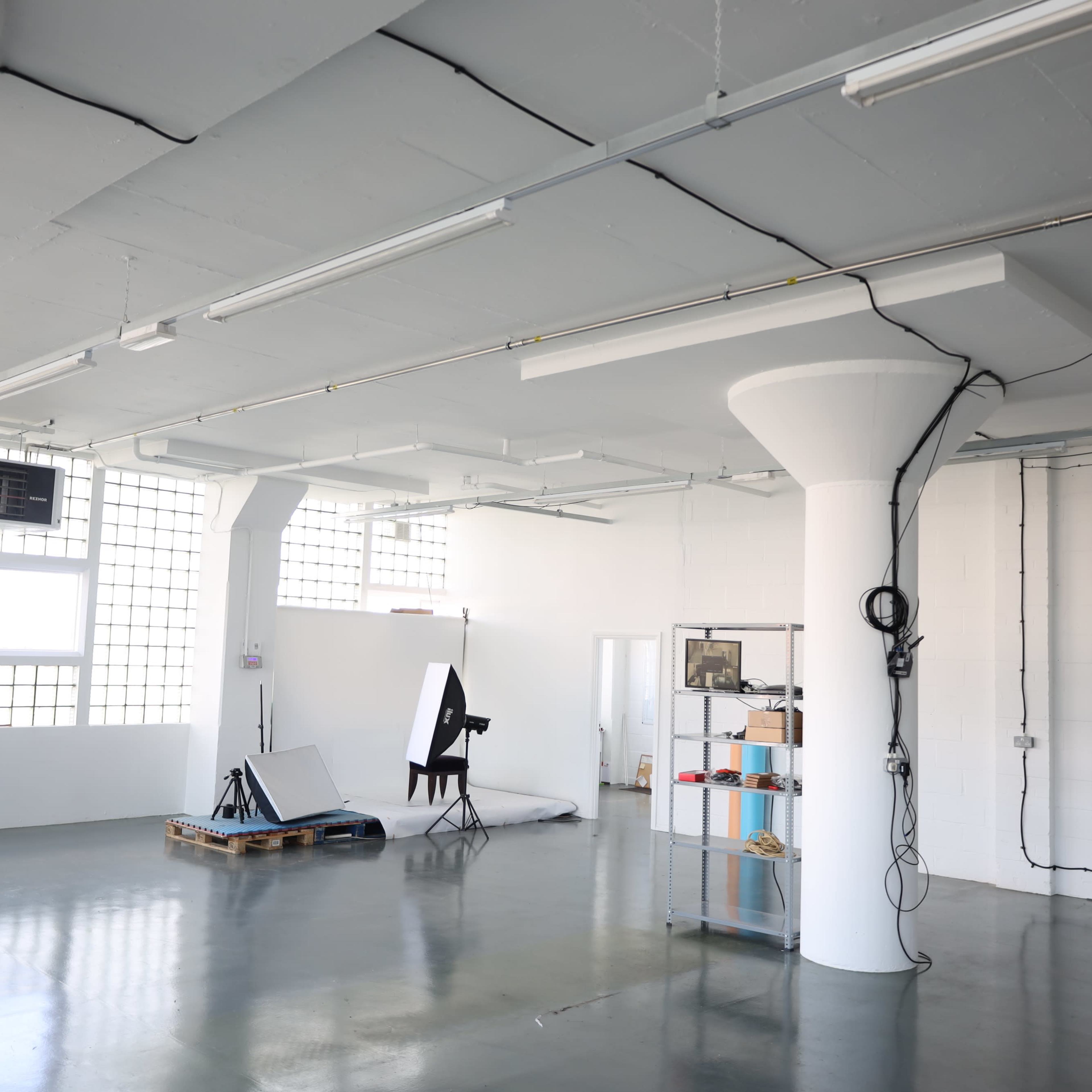 A spacious, empty studio with large windows, a single black chair, and shelves containing various items.