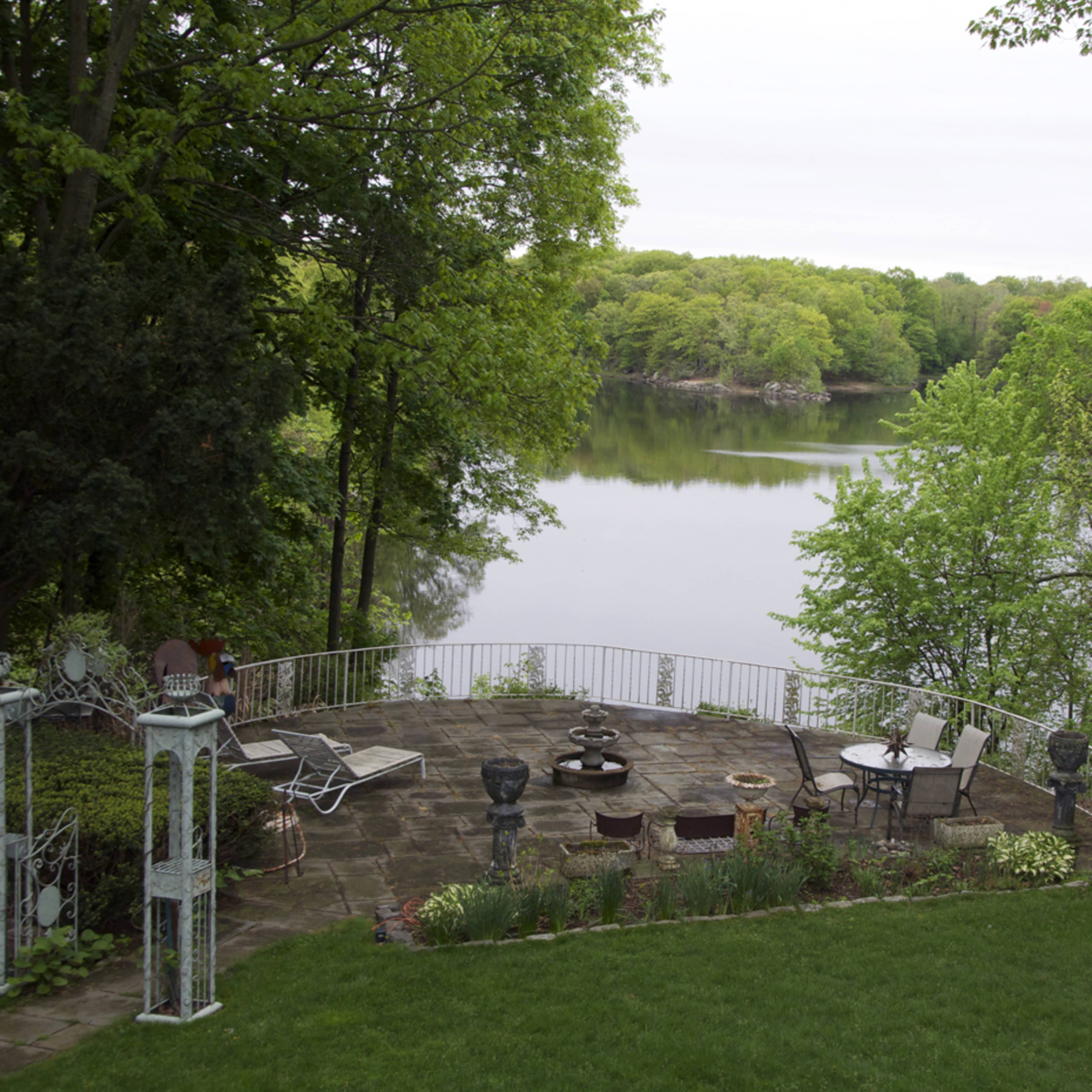 A stone patio with chairs and a fire pit overlooks a calm lake surrounded by trees.