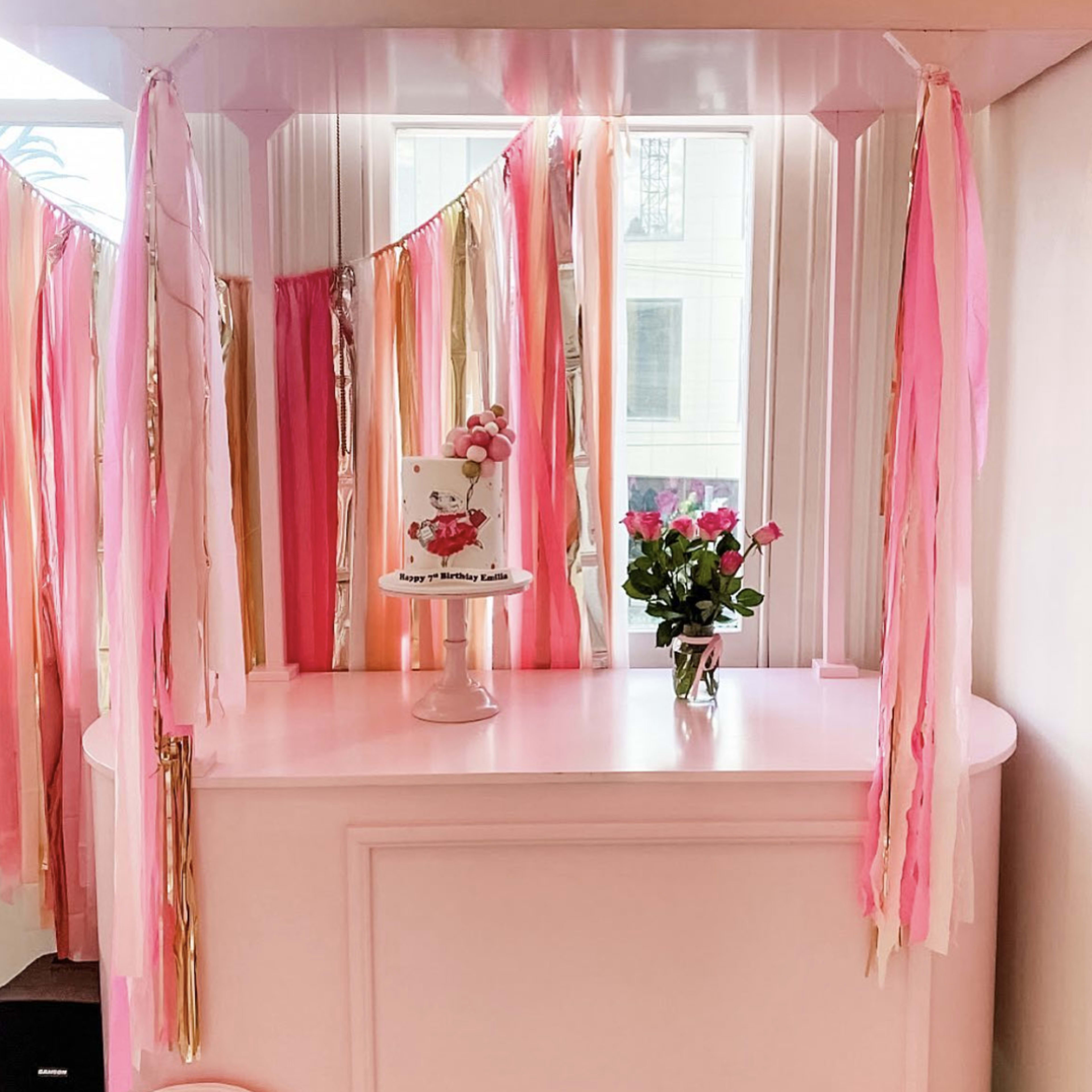 A decorated room featuring a pink and gold birthday cake on a white table, surrounded by colorful streamers and a vase of roses.