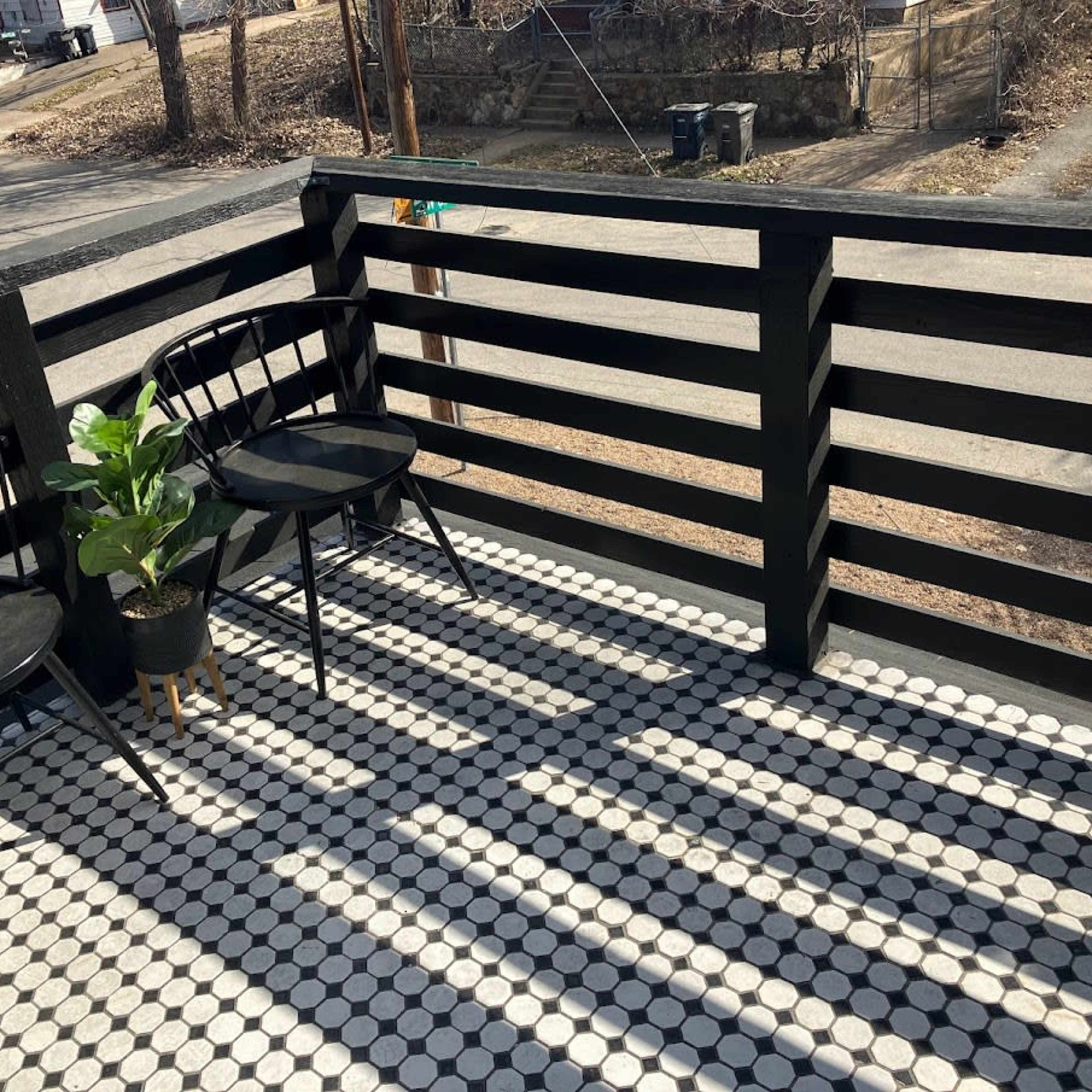 A balcony with two black chairs and a potted plant, featuring a patterned floor and shadows cast by the railing.