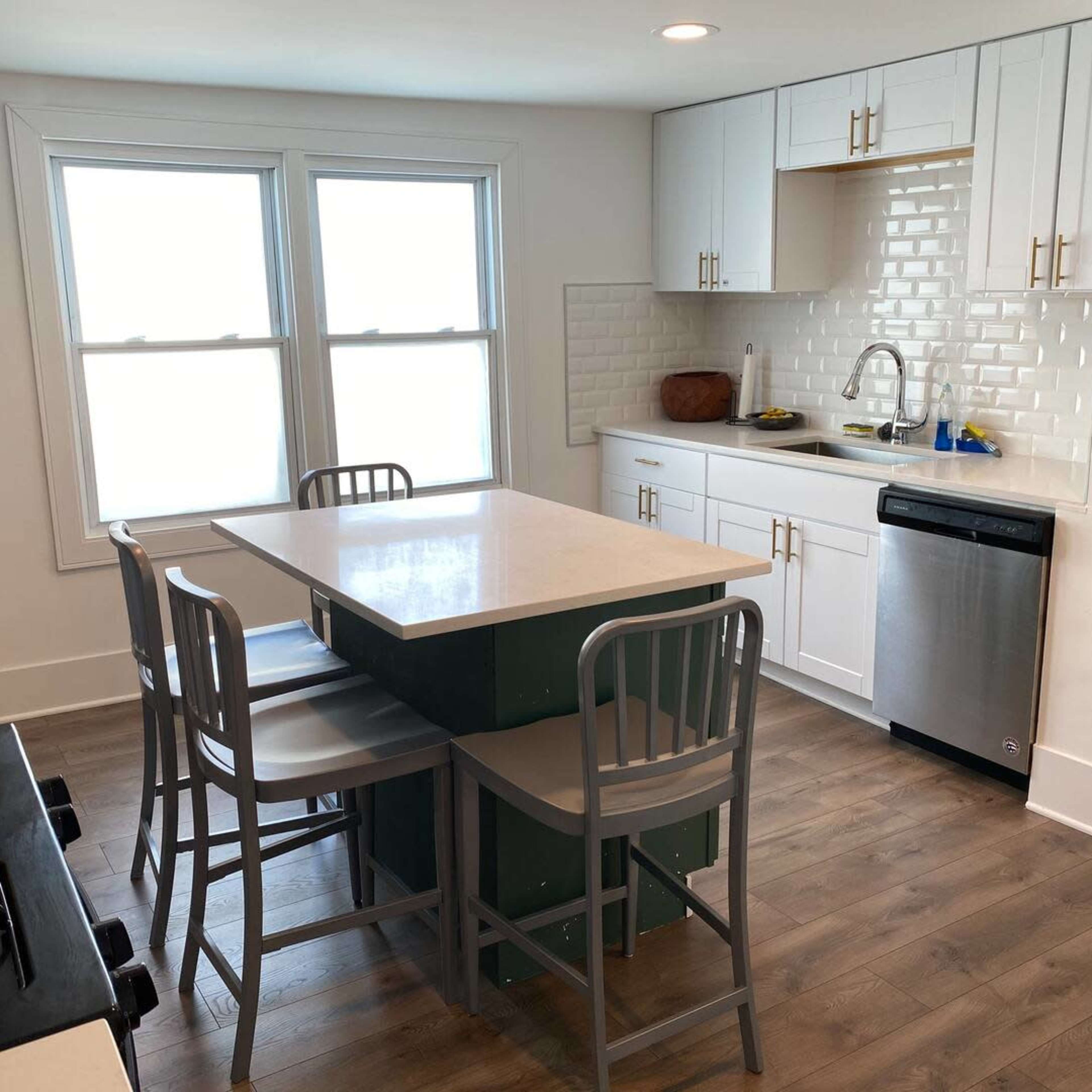 A modern kitchen features white cabinetry, a central island with seating, and large windows allowing natural light to enter.
