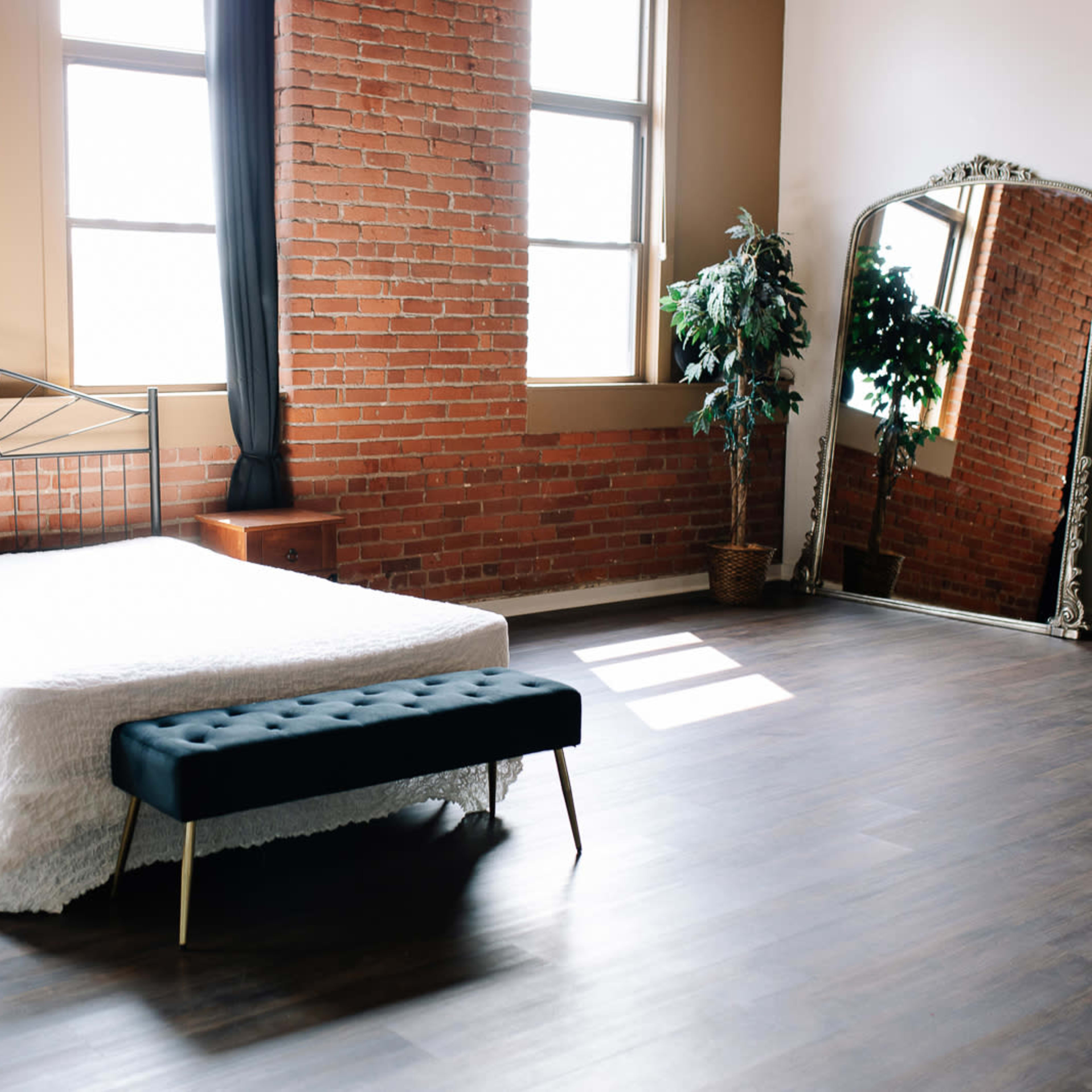 A minimalist bedroom features a metal bed frame with a white bedspread, a tufted black bench, and large windows alongside exposed brick walls.