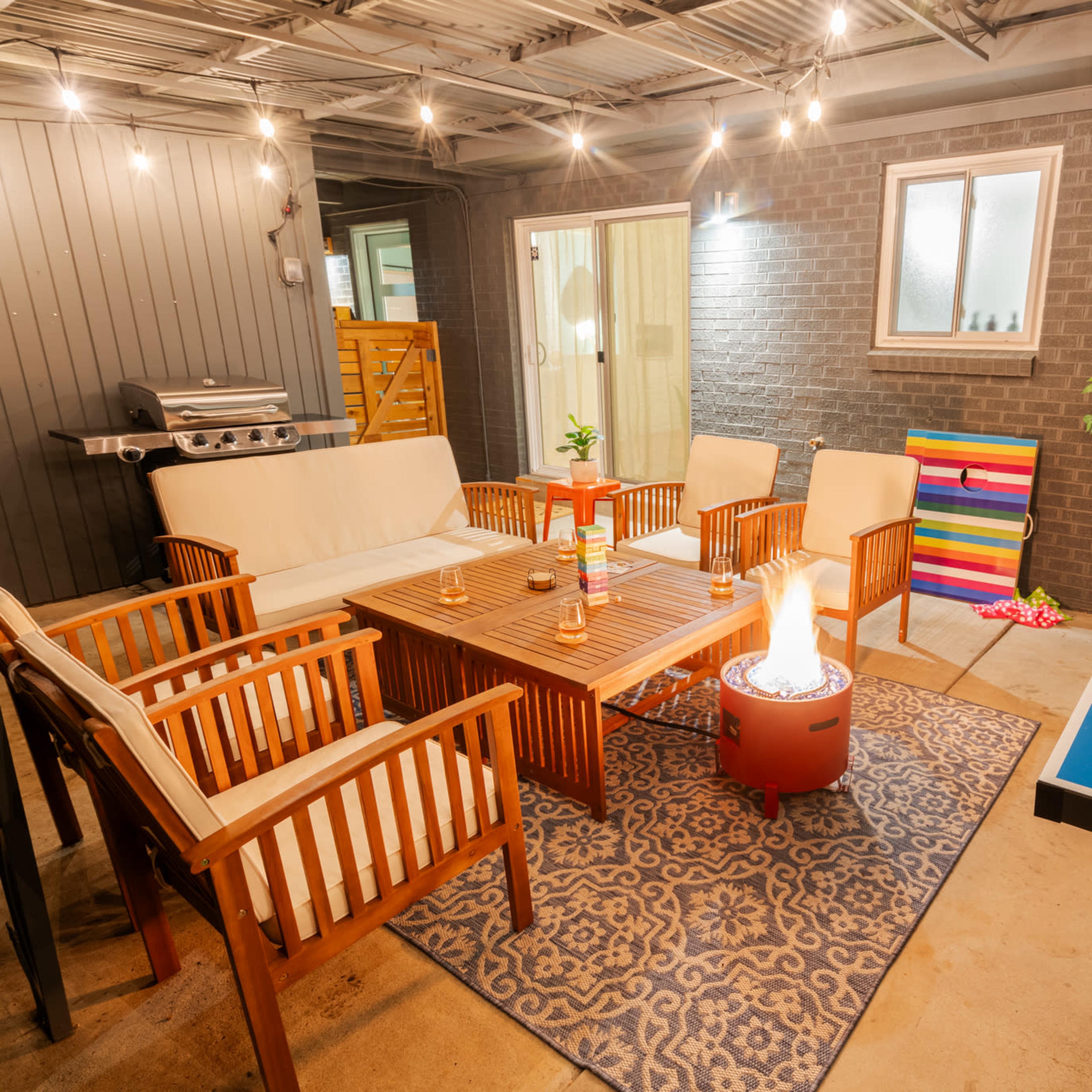 The scene features a patio area with seating around a table, a fire pit, a barbecue grill, and a ping pong table, all under string lights.
