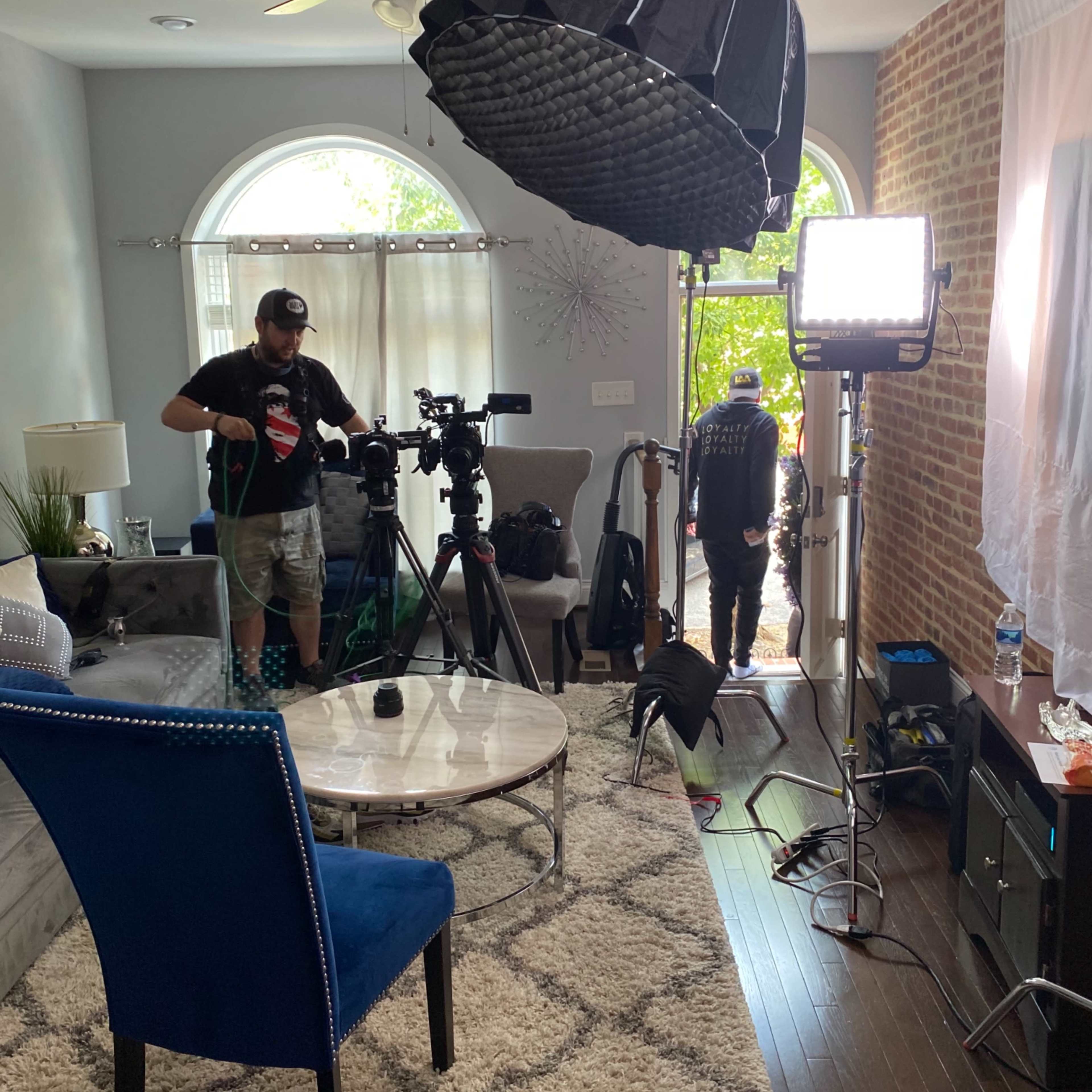A film crew sets up equipment in a well-lit living room with large windows and a patterned rug.