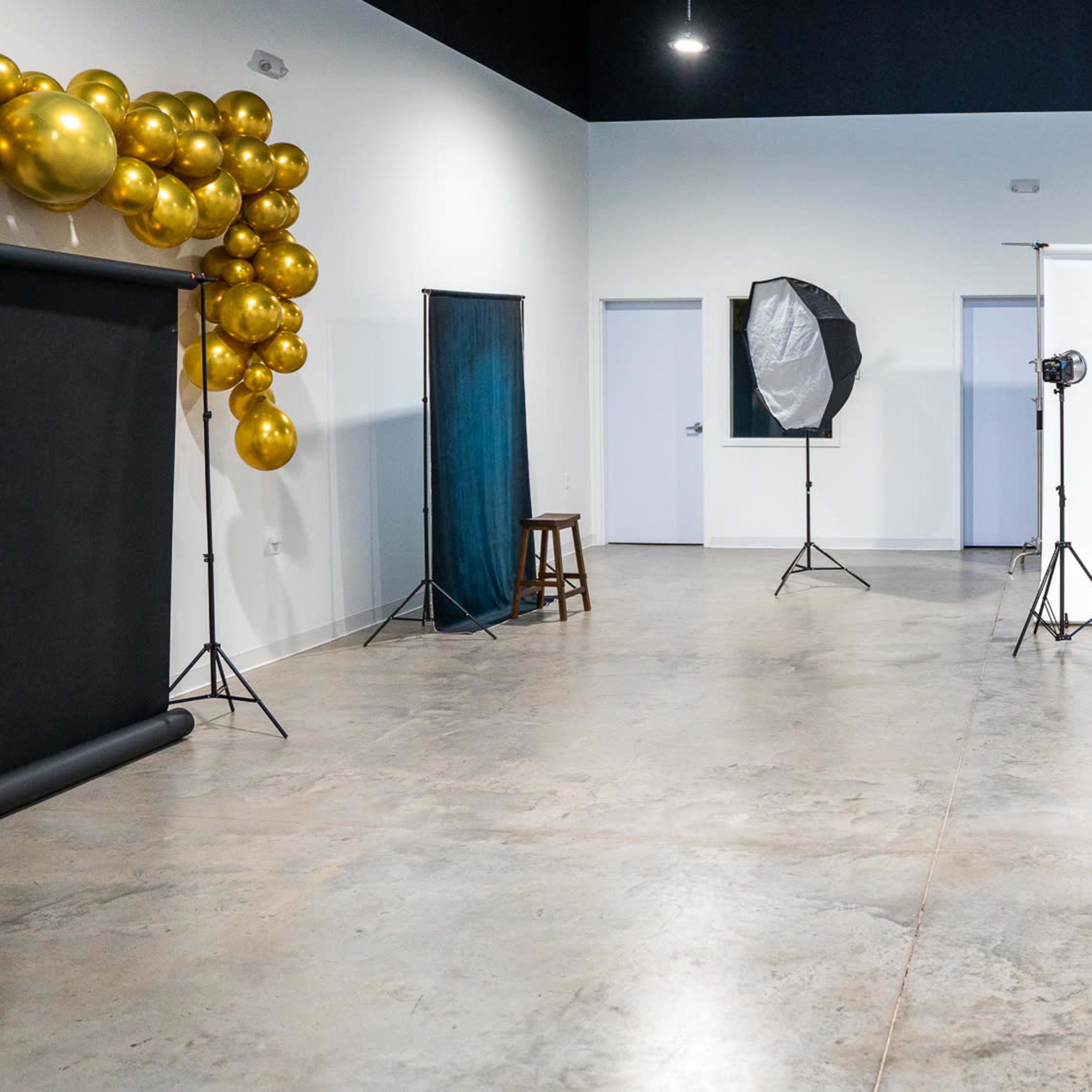 A spacious photography studio featuring a white backdrop, two light stands, a black backdrop setup, a stool, and a decorative arrangement of golden balloons on one wall.