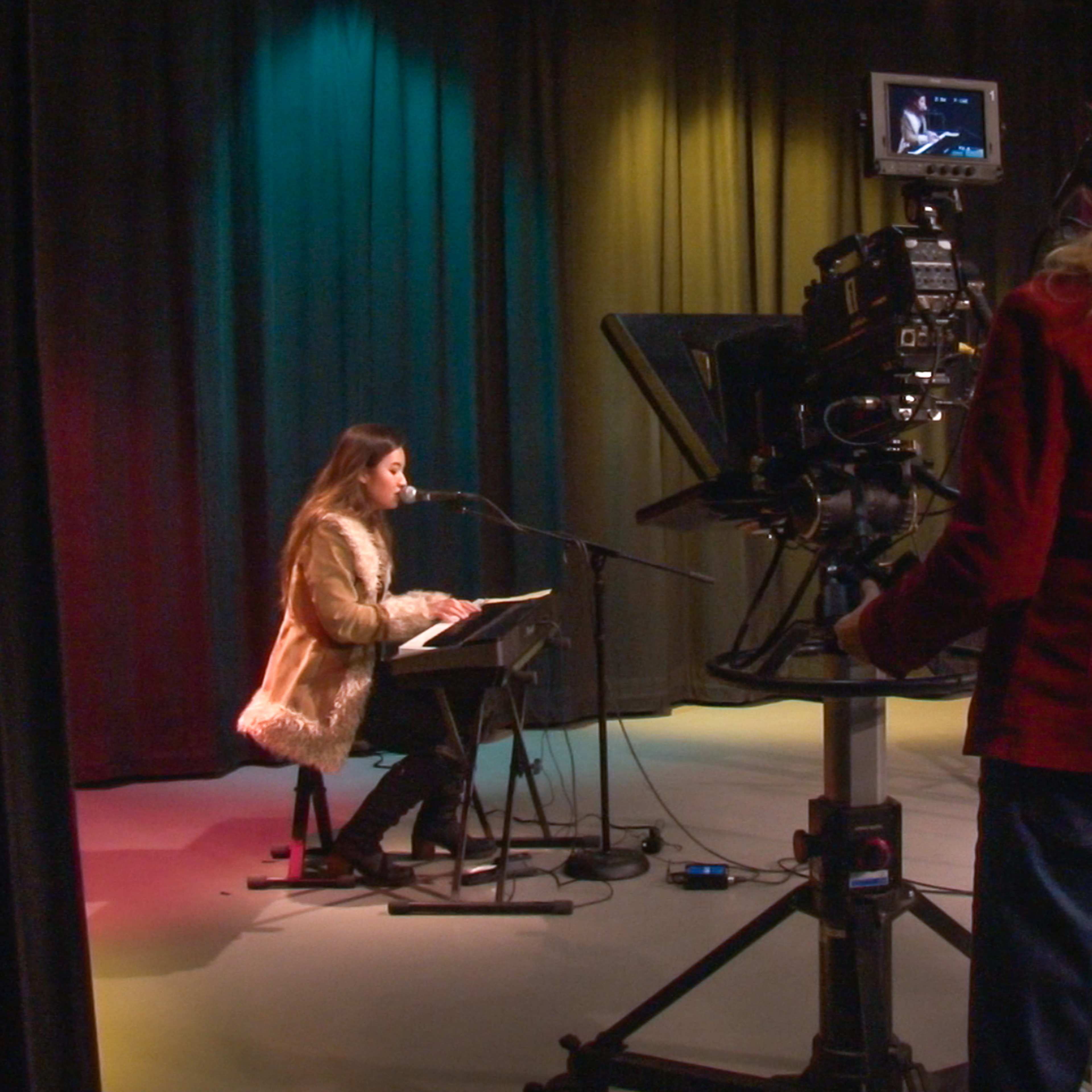 A woman in a faux fur coat performs at a microphone while another woman operates a video camera in a studio with colored backdrop lights.