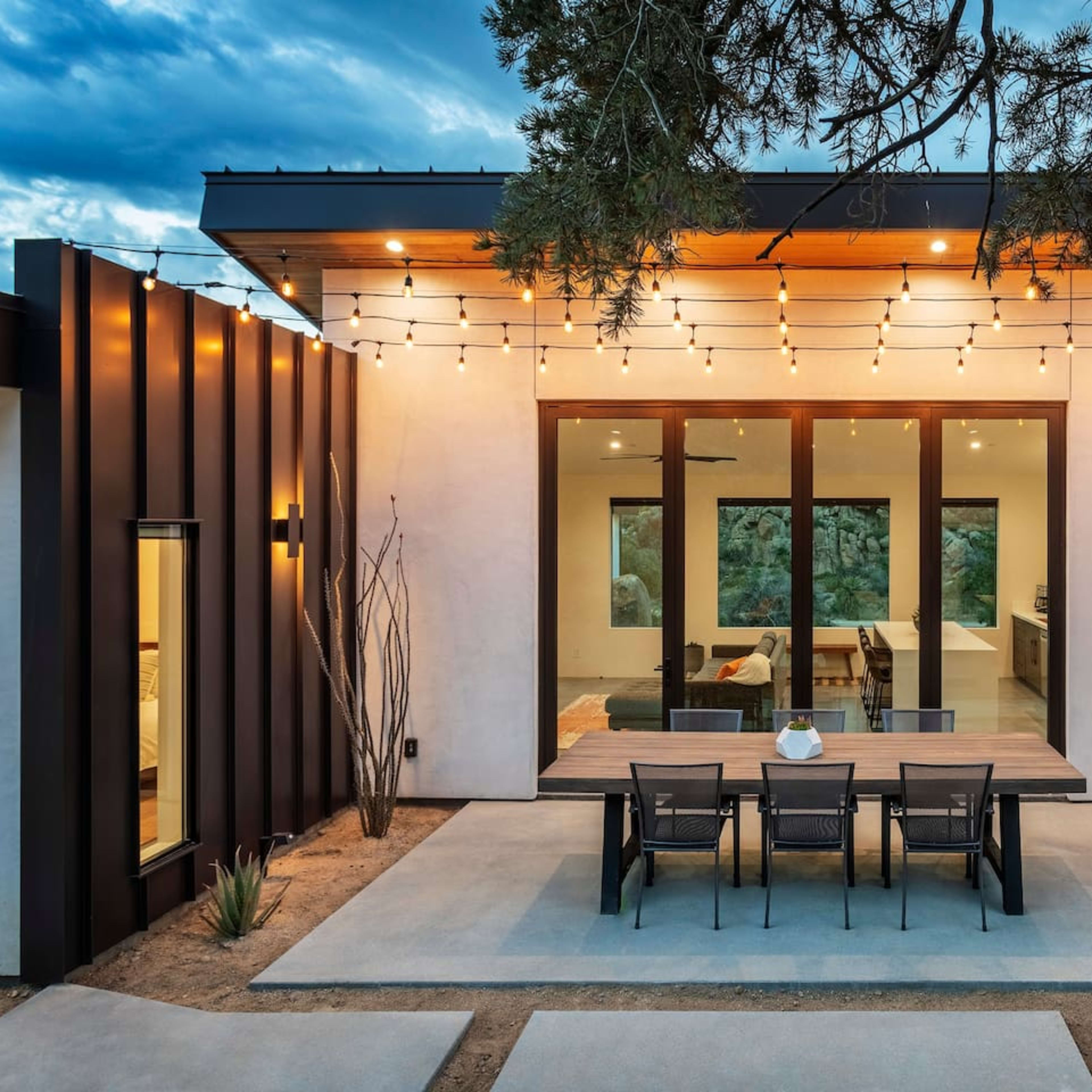 A modern home with a patio area featuring a large table and string lights, surrounded by desert landscaping.
