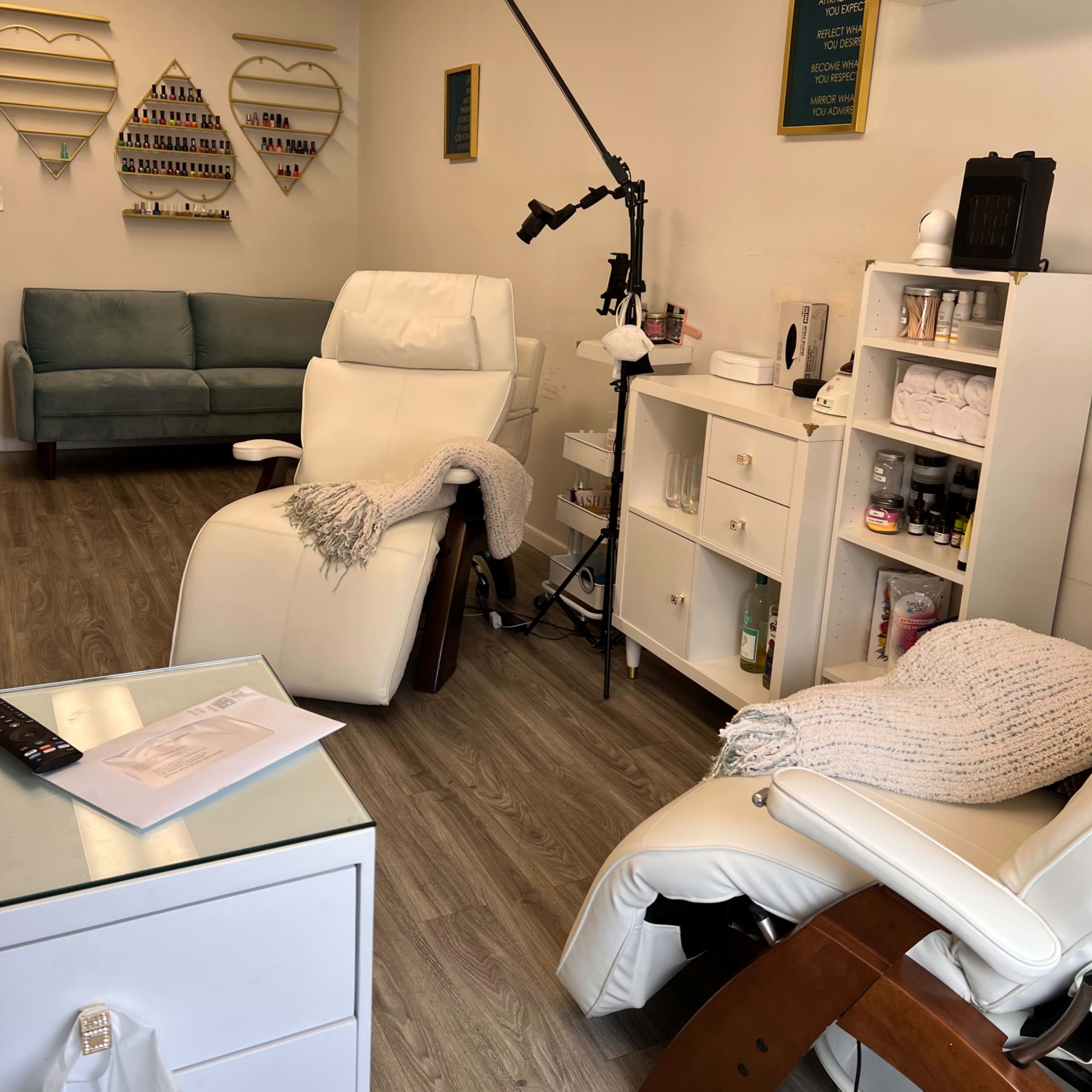 A nail salon interior featuring two reclining chairs, a manicure station, a couch, and various nail polish displays on the walls.