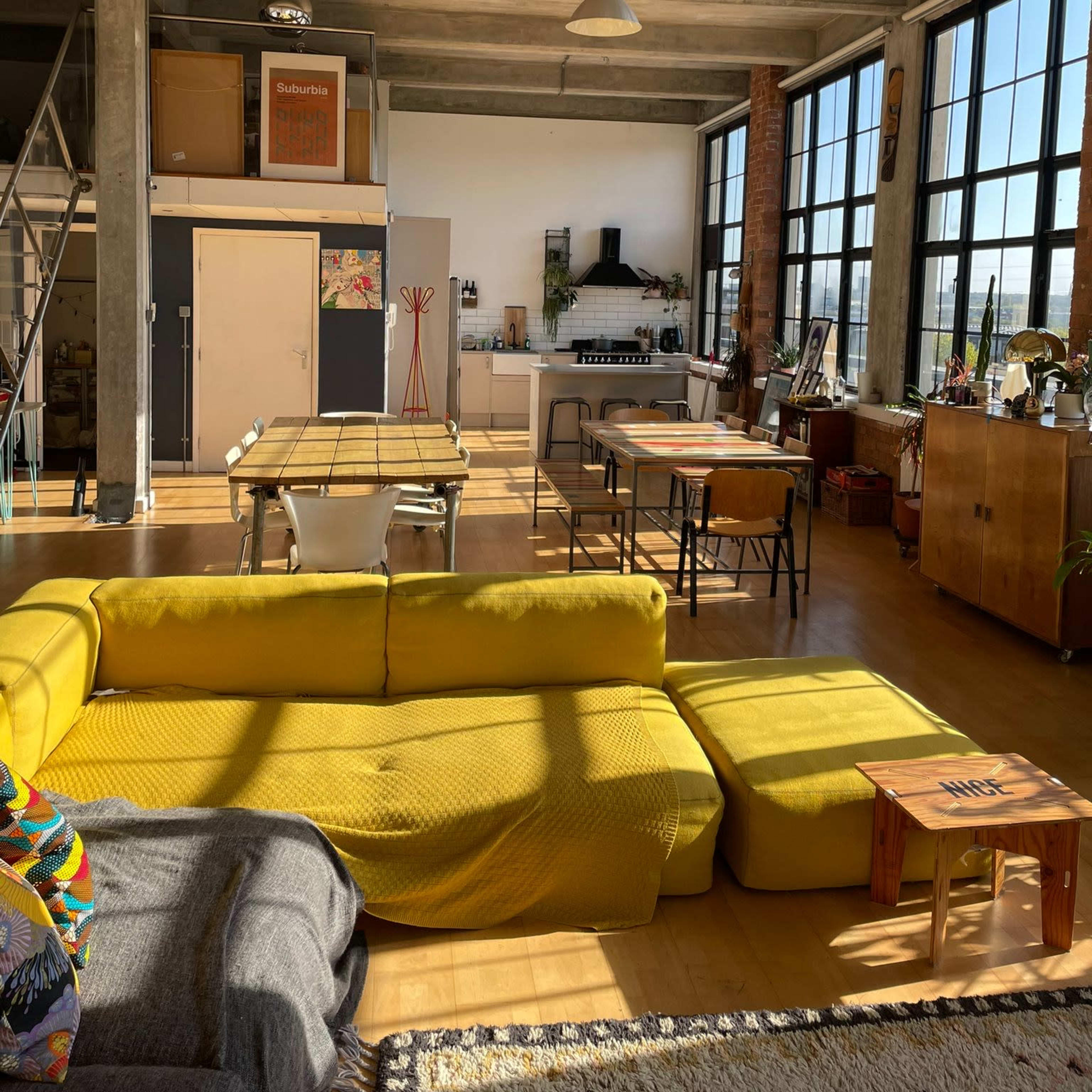 A spacious, modern loft-style living area with large windows, a yellow sectional sofa, a dining table, and a kitchen in the background.