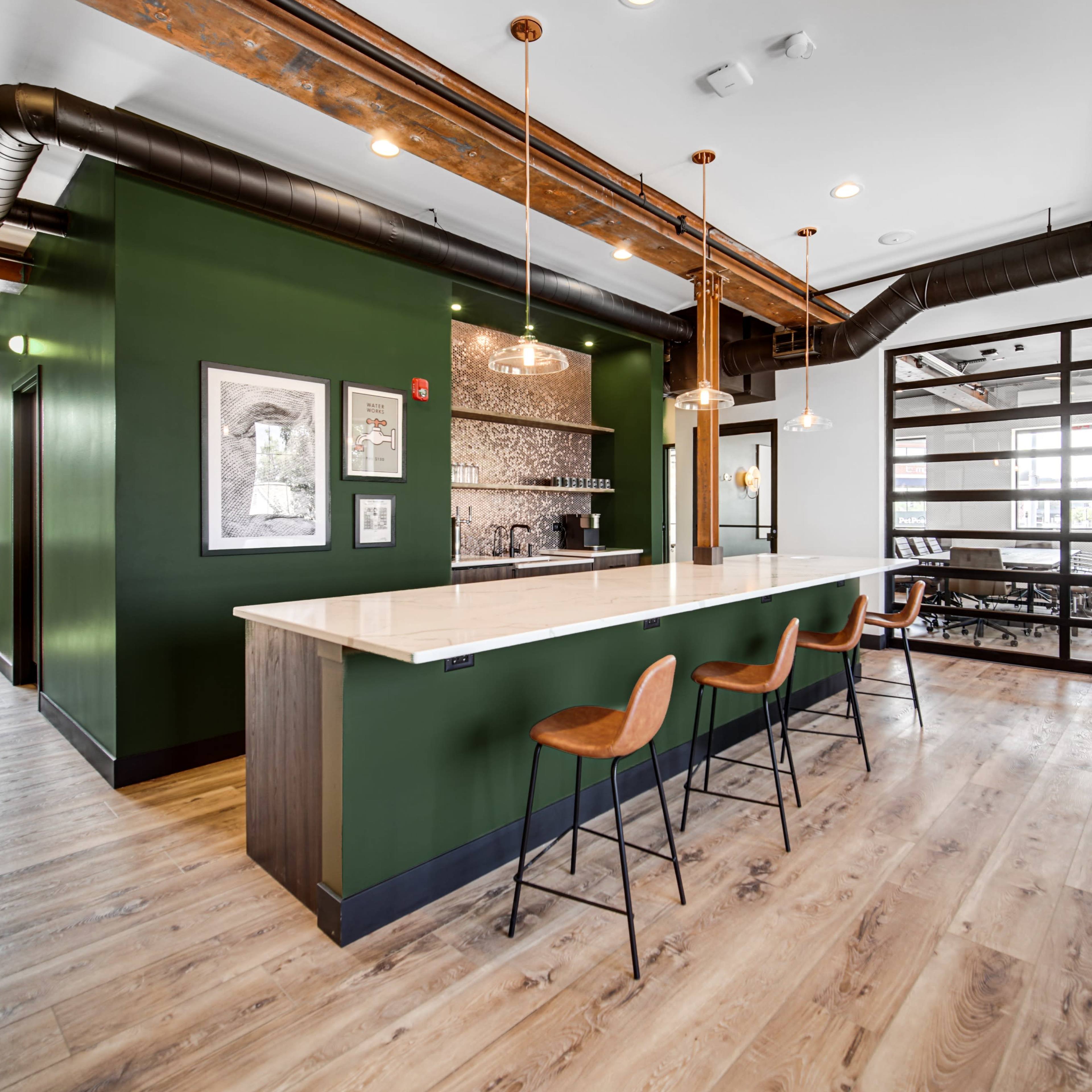 A modern kitchen area with a long countertop and wooden bar stools, featuring green walls and exposed ceiling beams.