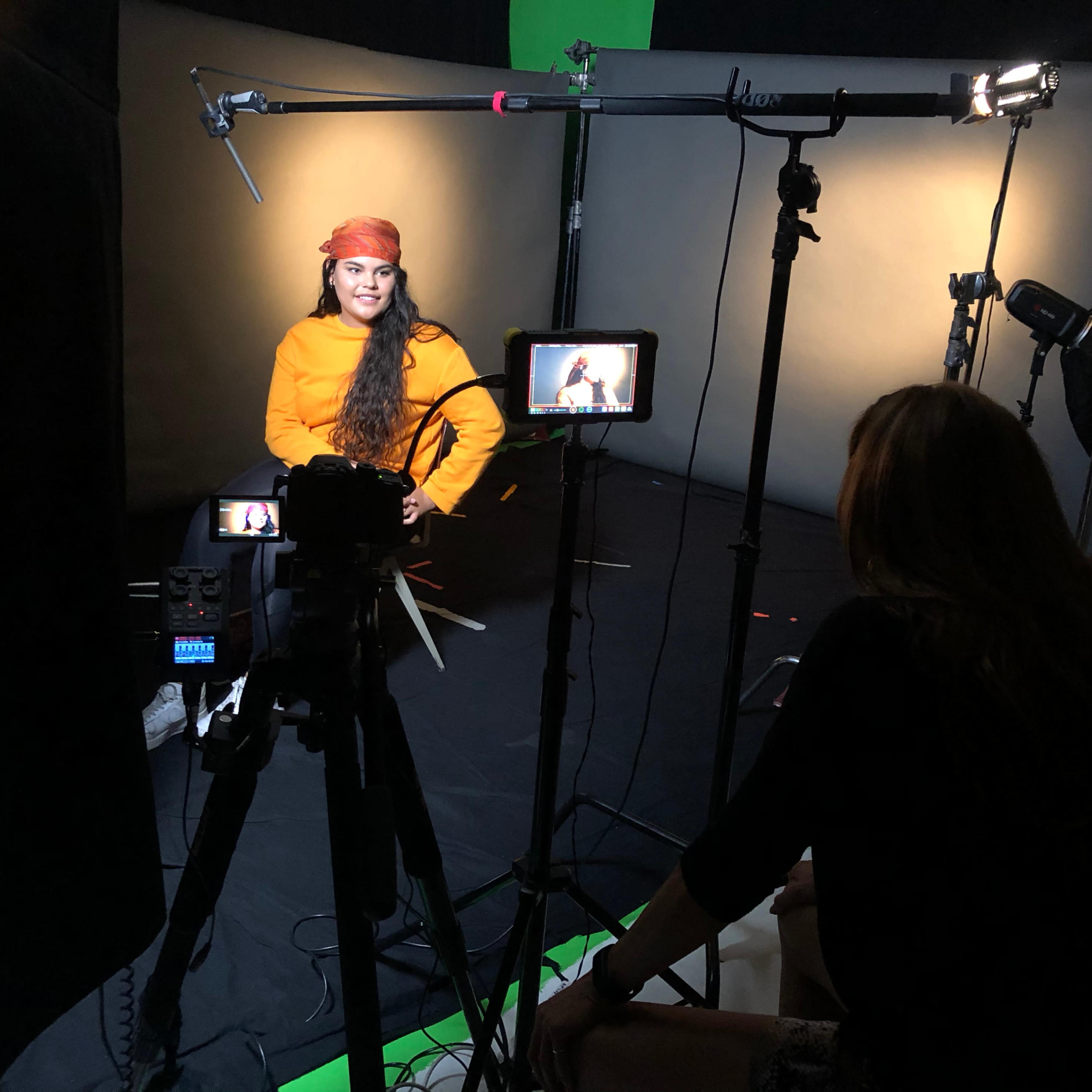 A woman in a bright orange shirt and a headband is seated in front of a camera setup, while another individual adjusts equipment nearby.