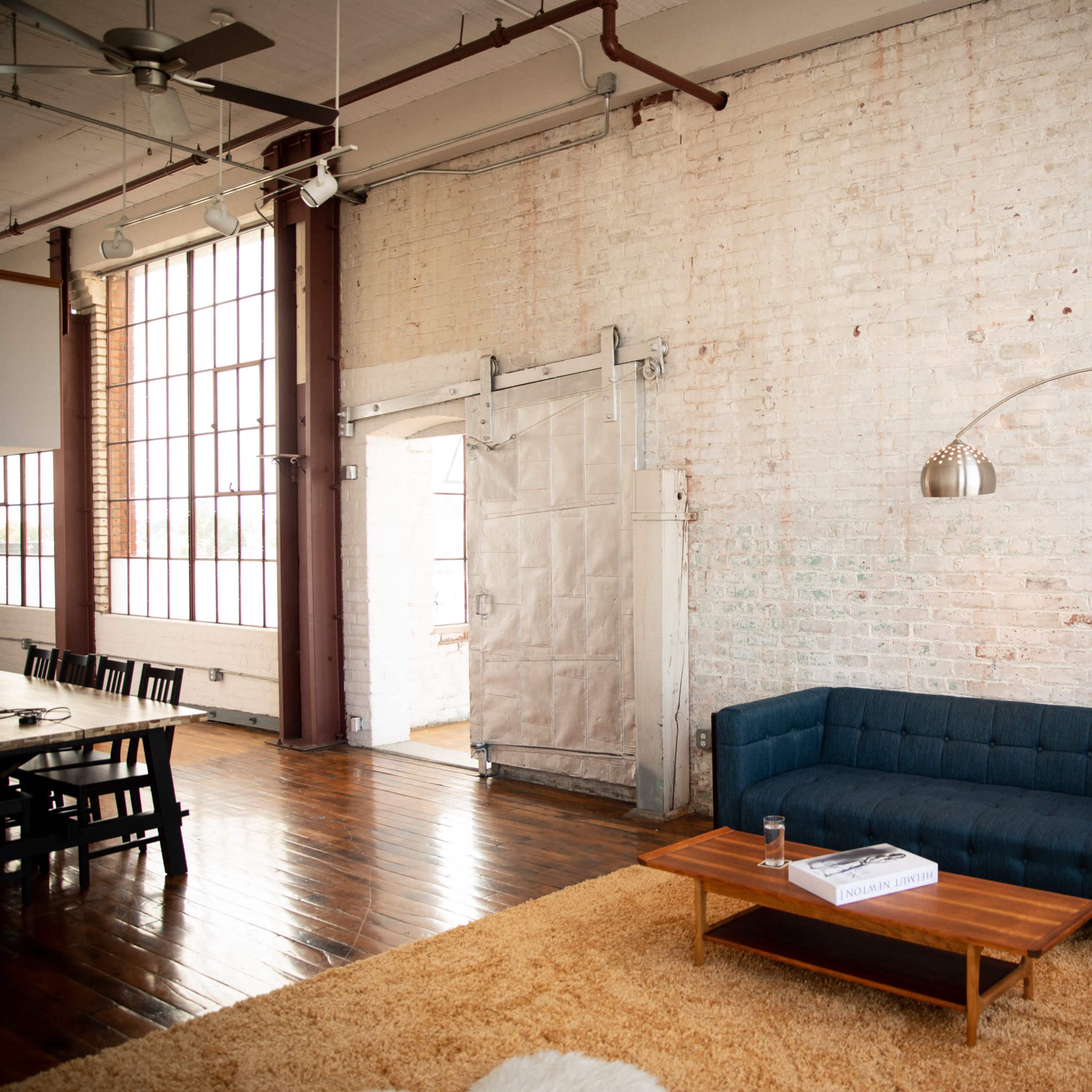A modern industrial living space featuring a blue sofa, an arc floor lamp, and a wooden coffee table on a textured rug against a brick wall.