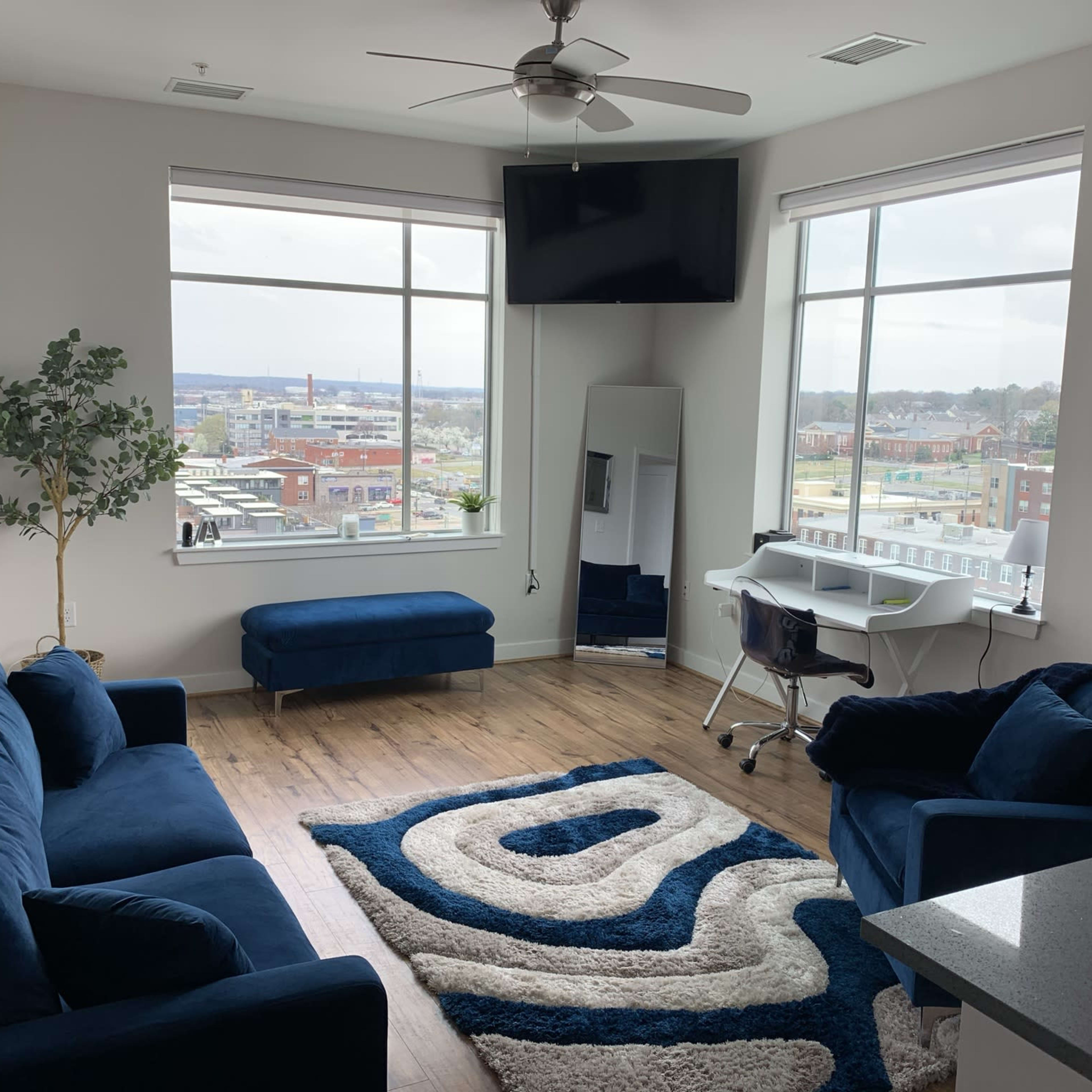 A modern living room with two blue sofas, a mirror, a desk with a chair, and large windows overlooking an urban landscape.