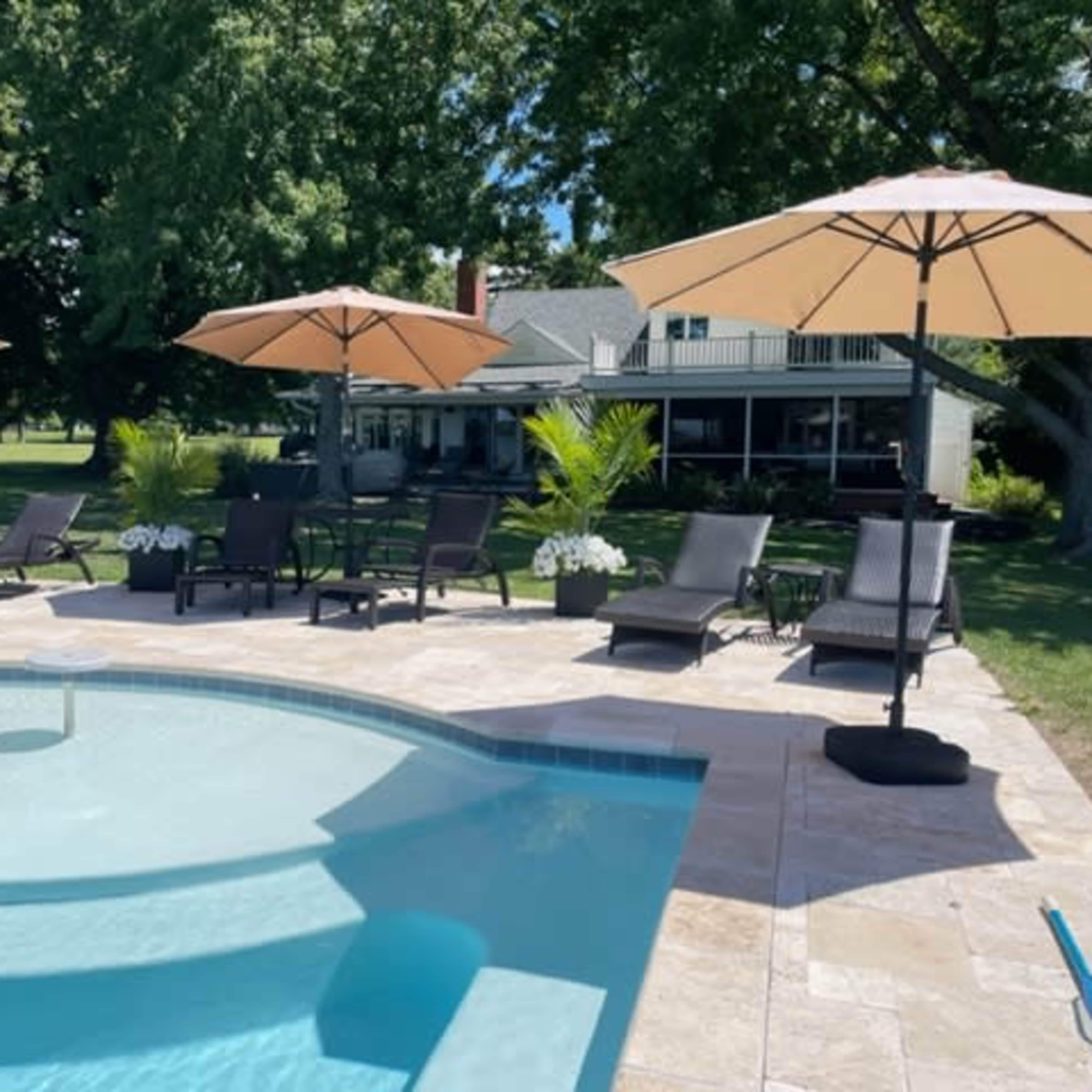 A swimming pool area with lounge chairs and umbrellas, set in a grassy yard with a house in the background.