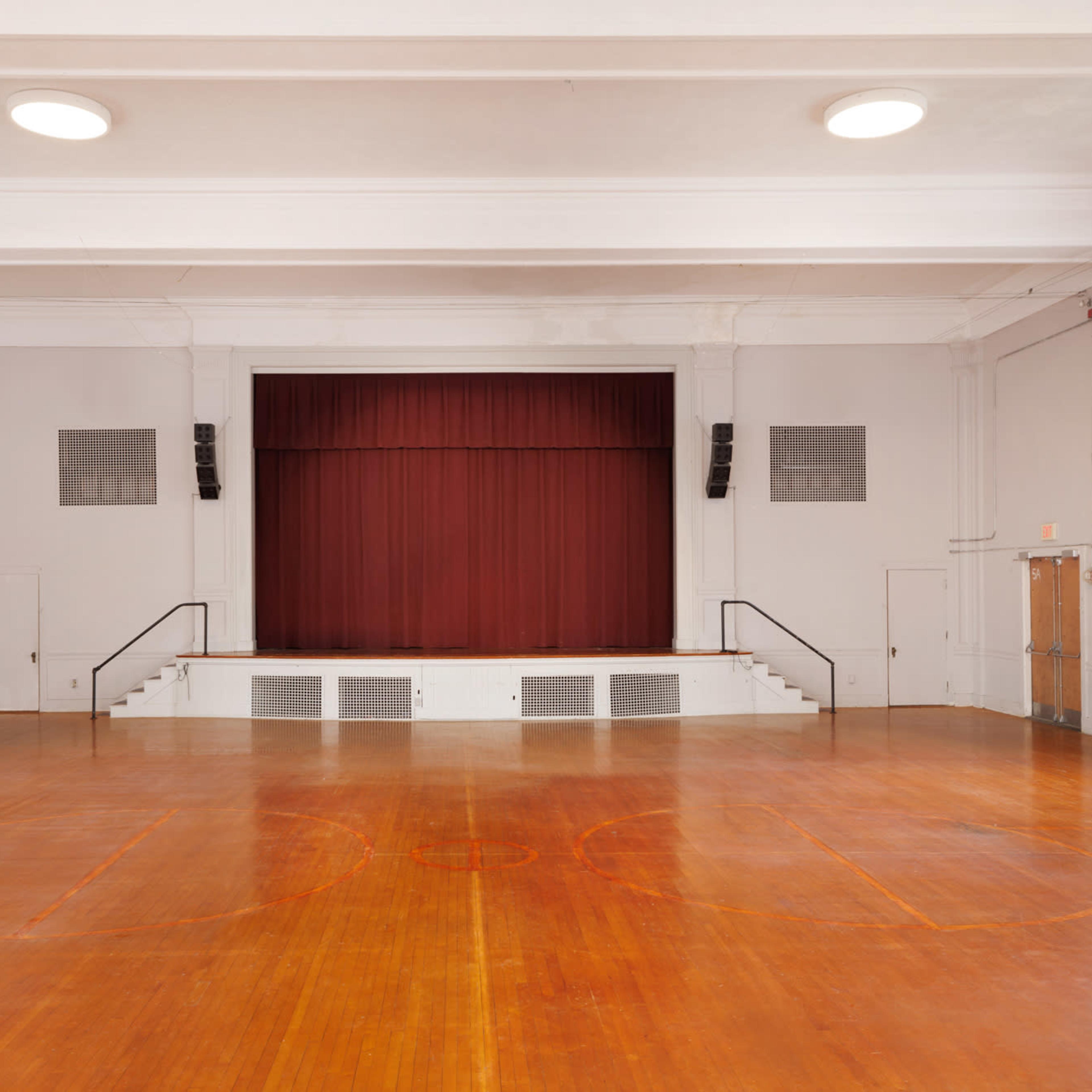 An empty gymnasium with a wooden floor and a stage at the front covered by a red curtain.