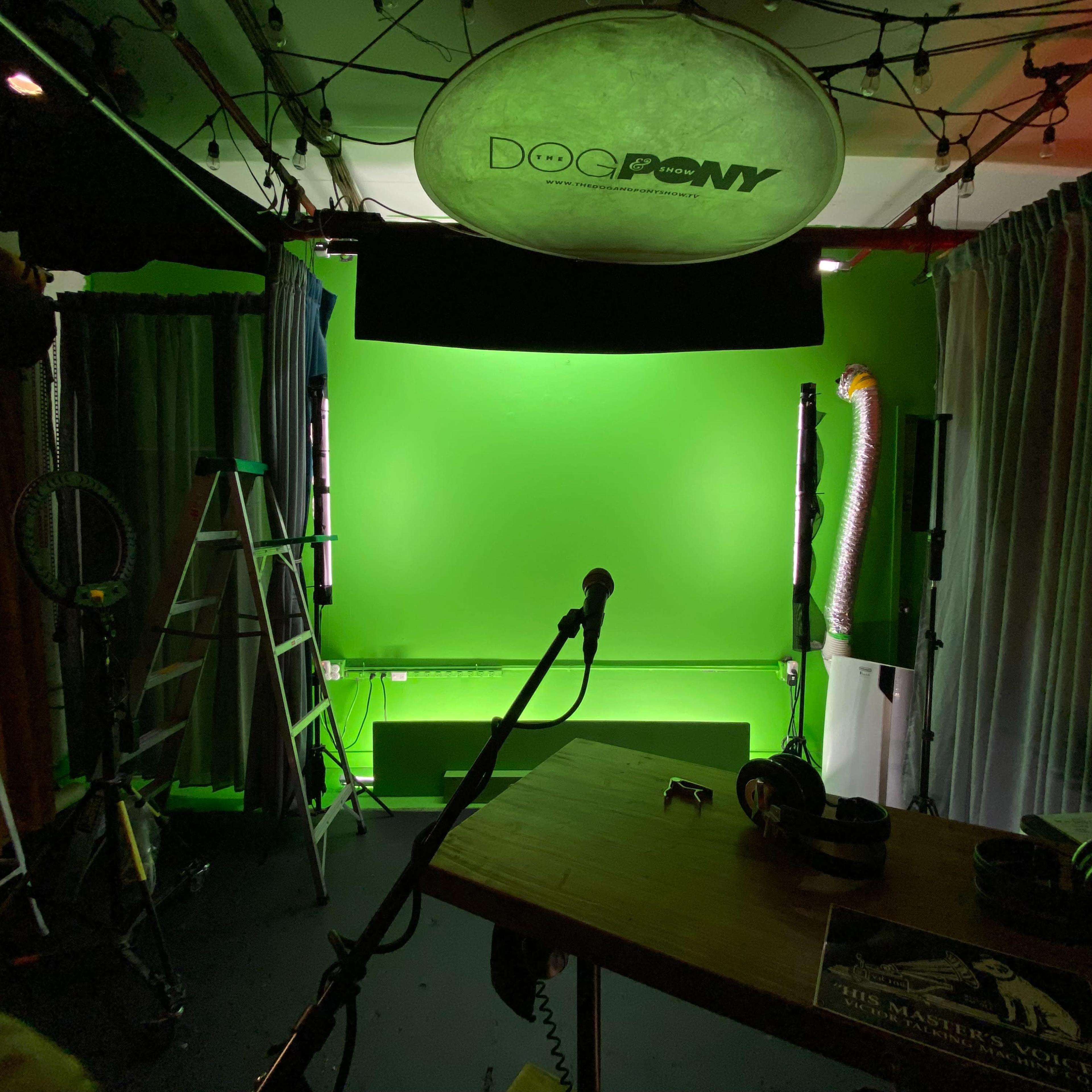 A studio space with a green screen backdrop, a microphone on a stand, a table with headphones, and a ladder in the background.