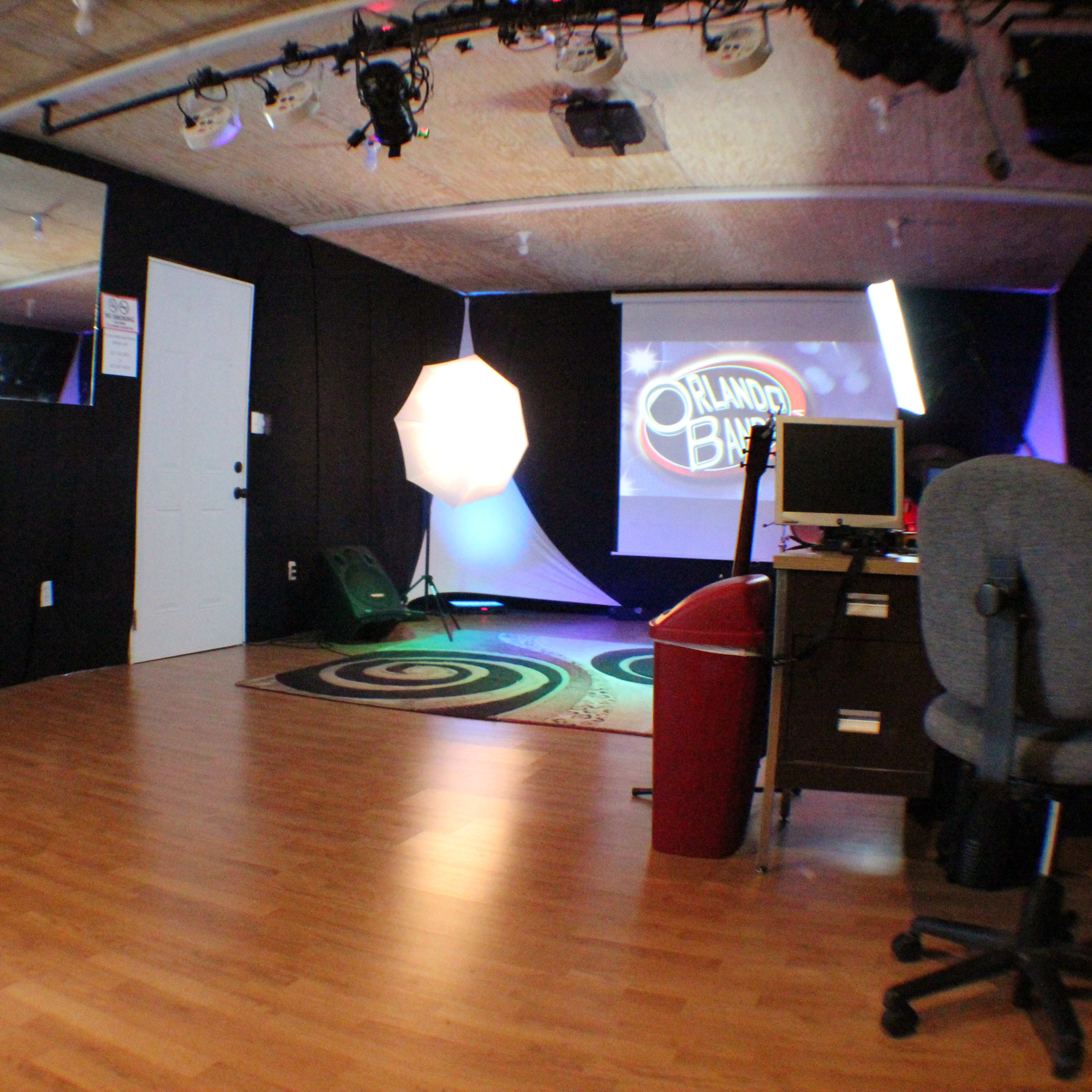 A small studio space with a wooden floor, a gray office chair, a desk with a computer, and lighting equipment set up in front of a backdrop featuring the words "Project B."