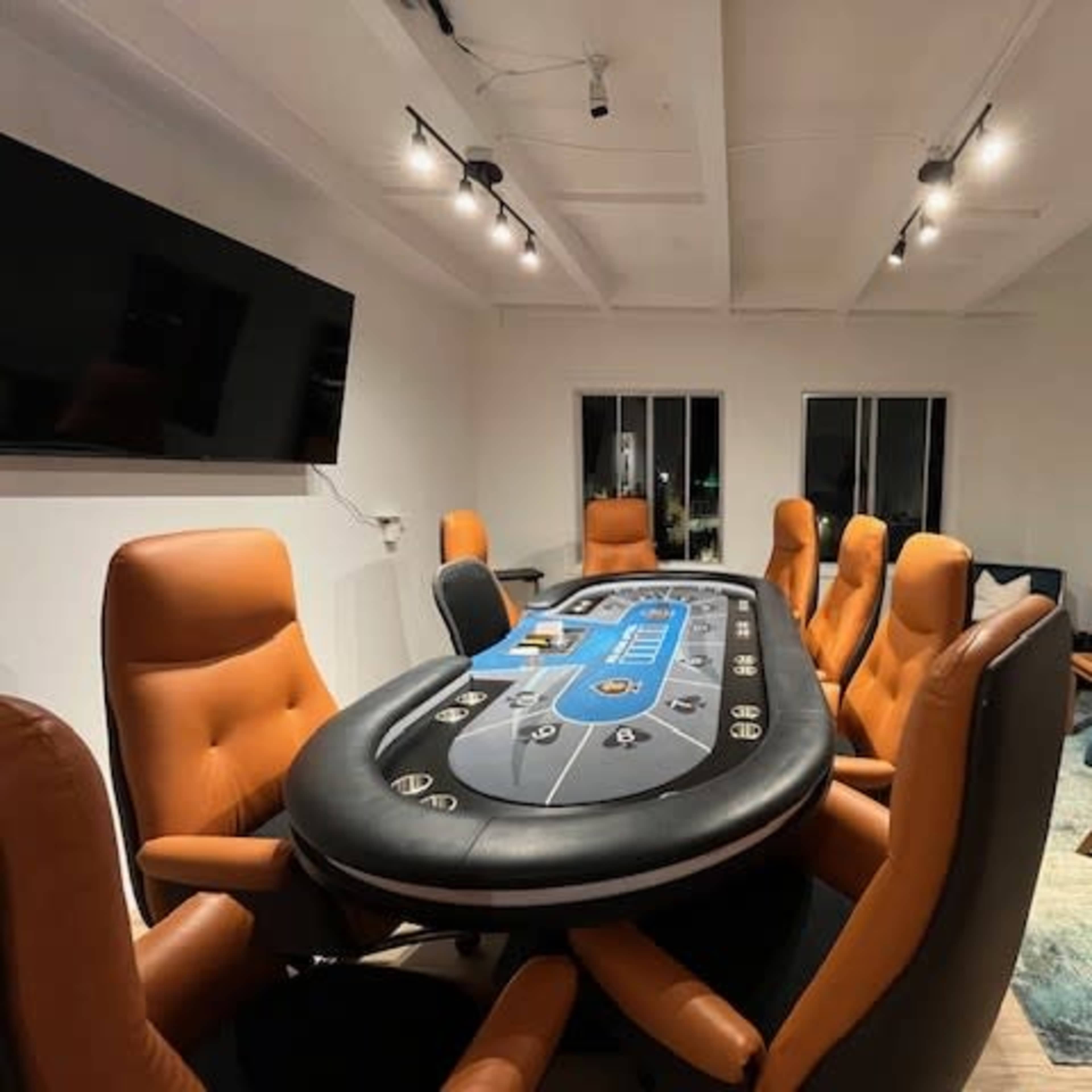 A poker table with eight brown leather chairs is set up in a brightly lit room featuring a large abstract painting and a blue upholstered chair.