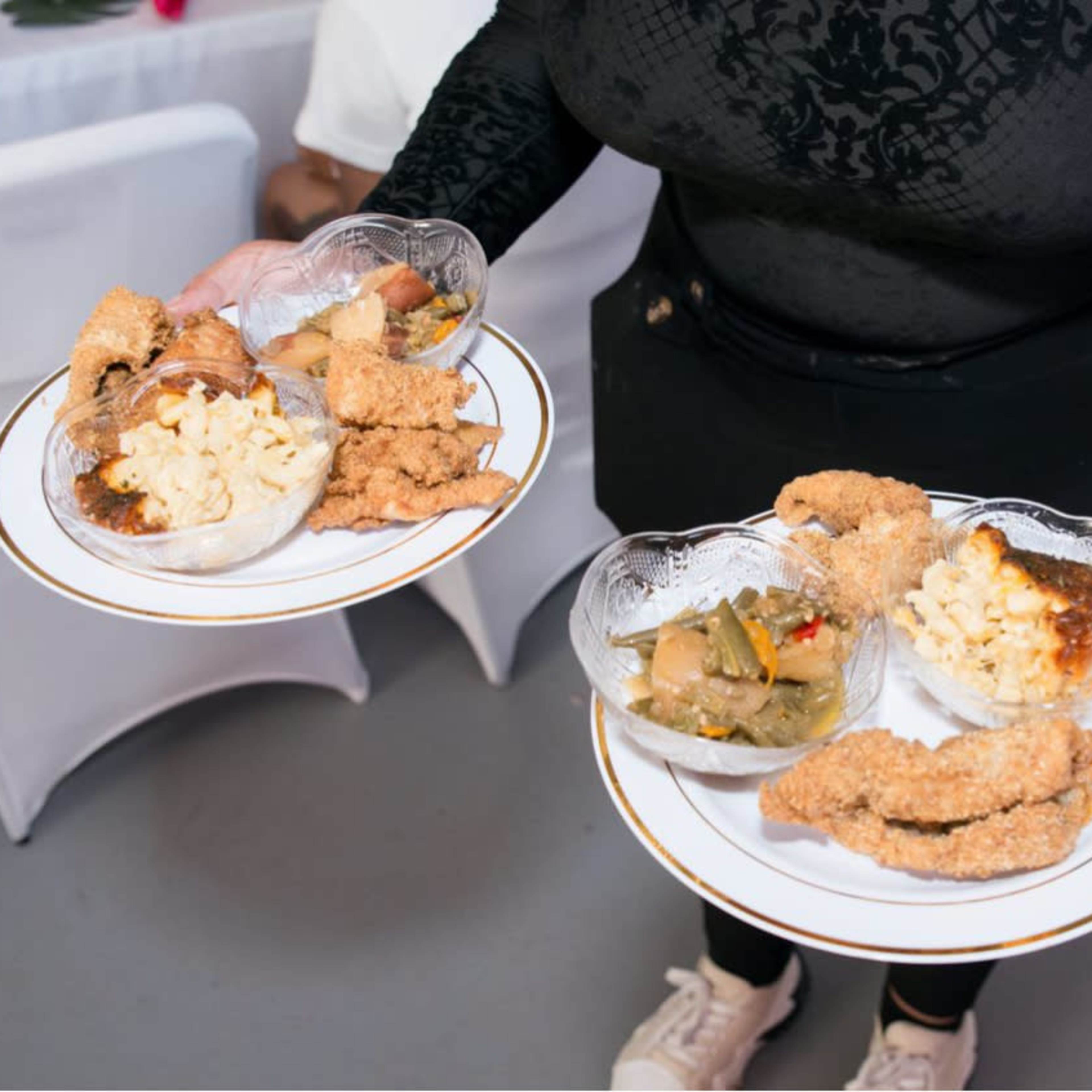 A person is holding two plates filled with fried chicken, macaroni and cheese, and a vegetable dish.
