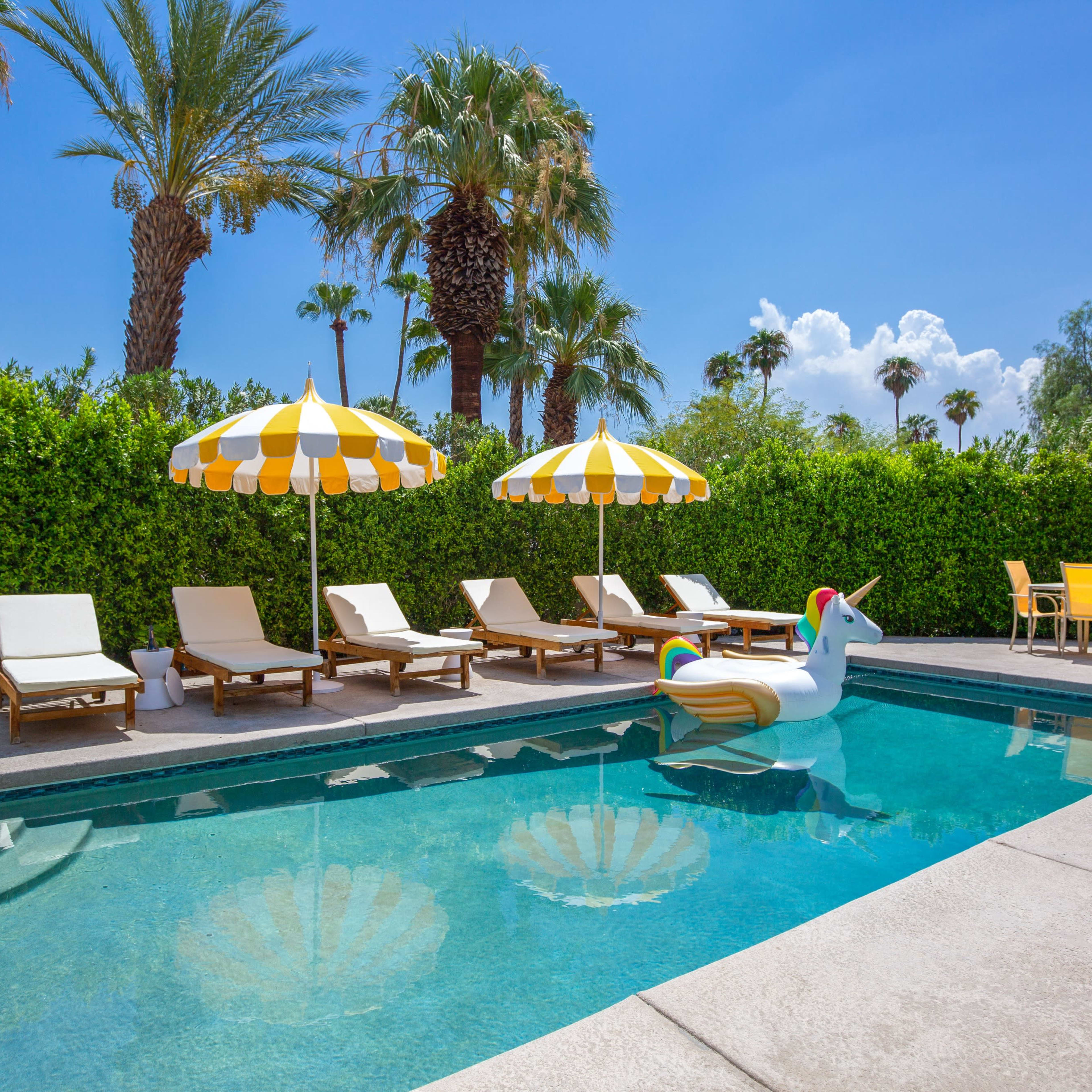 A swimming pool surrounded by lounge chairs, yellow and white umbrellas, and a large inflatable unicorn in a tropical setting.