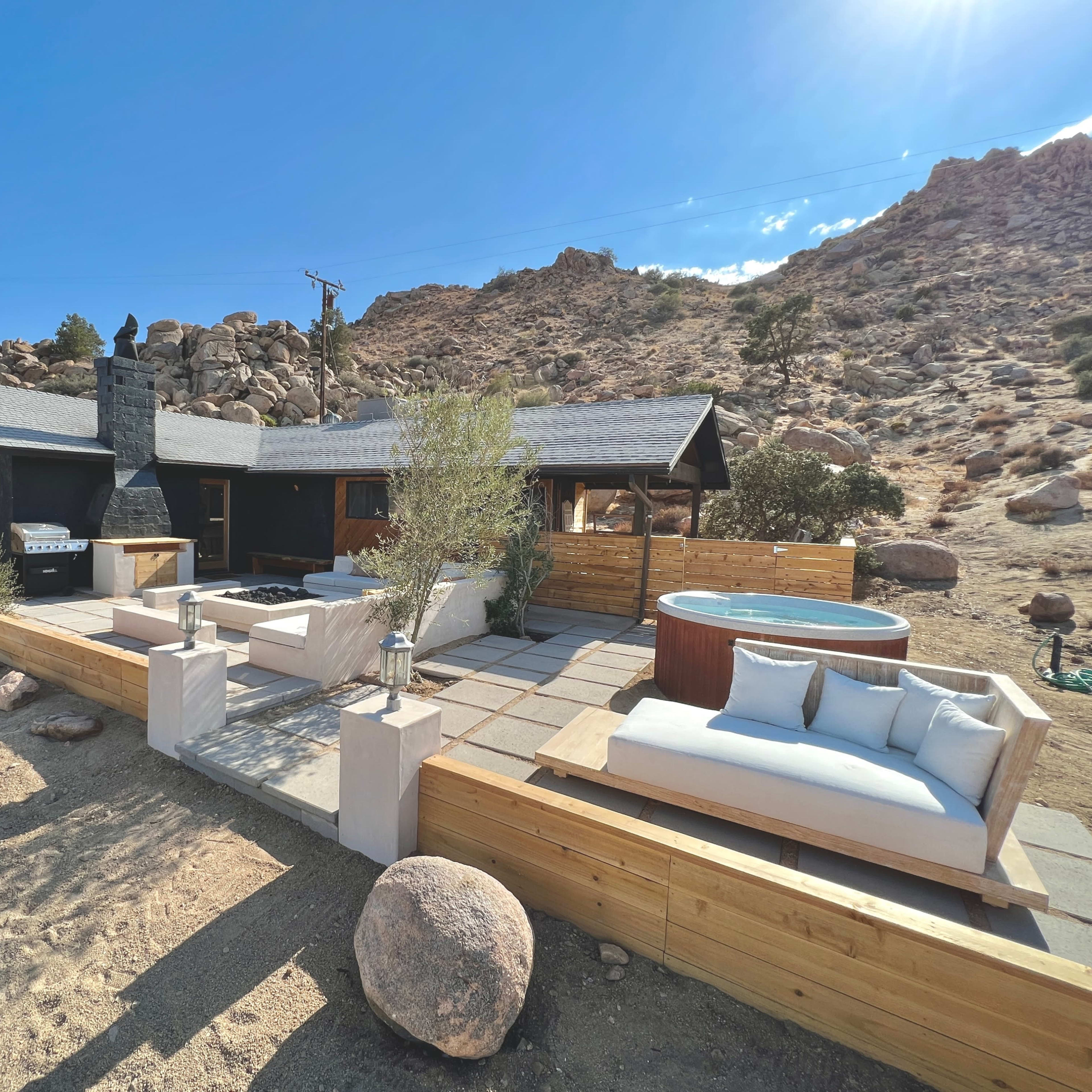 A modern outdoor patio area with a hot tub, seating, and a charcoal grill, surrounded by rocky terrain and mountains.