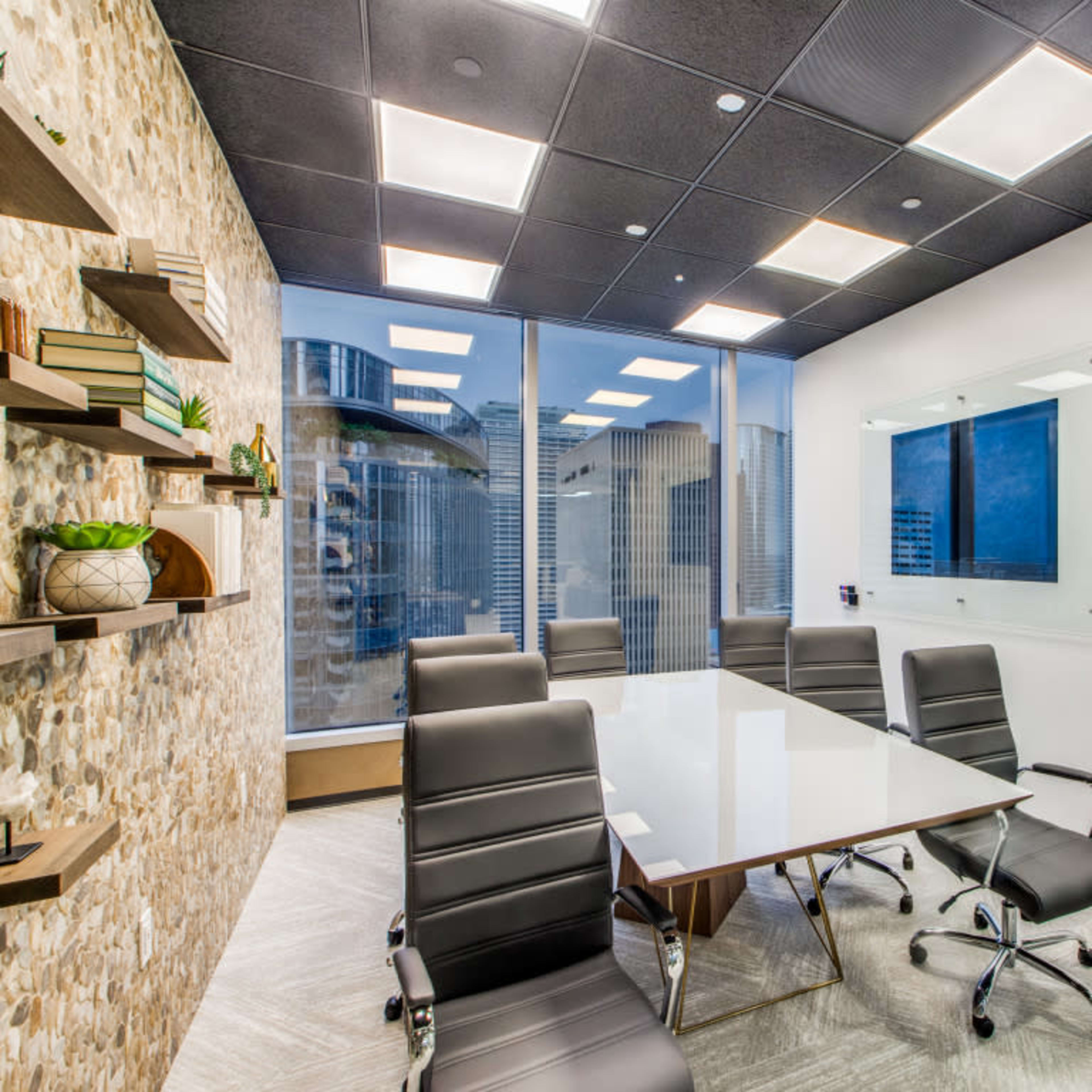 A modern conference room with a glass wall, a large white table, and black leather chairs arranged around it, overlooking a city skyline.