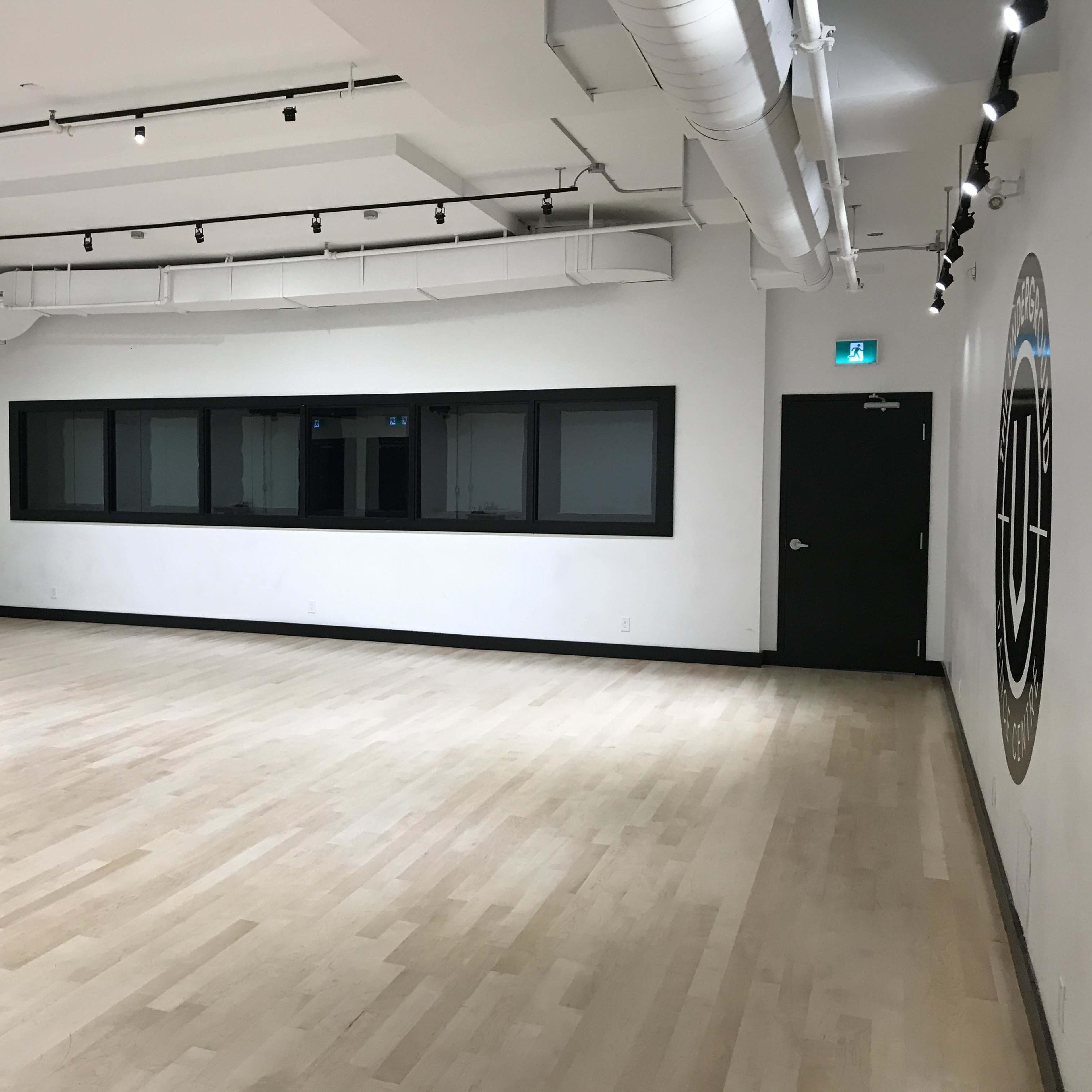 A spacious dance studio with wooden flooring, large windows, and a mirror along one wall.