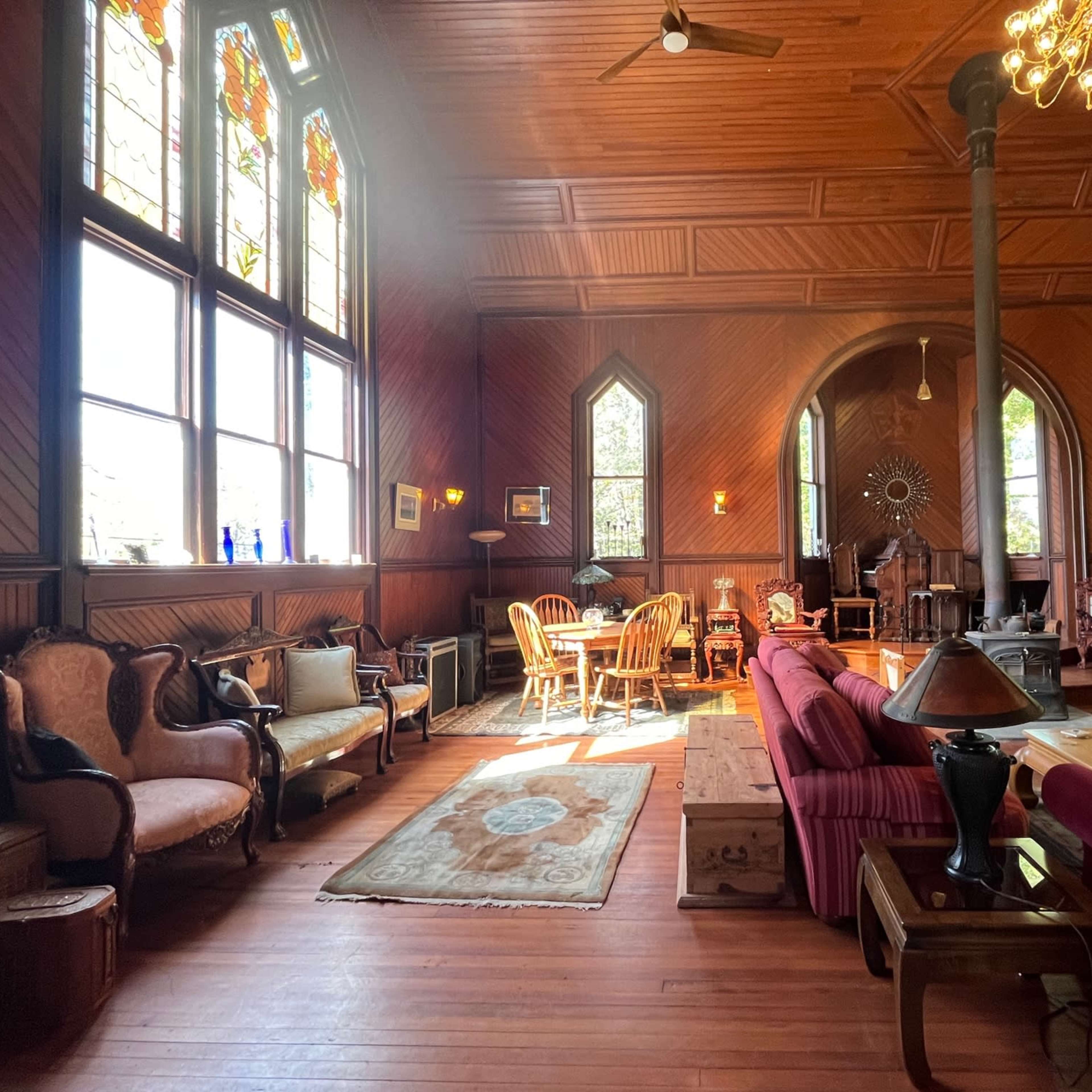 The spacious room features wooden walls, large stained glass windows, and a mix of vintage furniture arranged around a central seating area.
