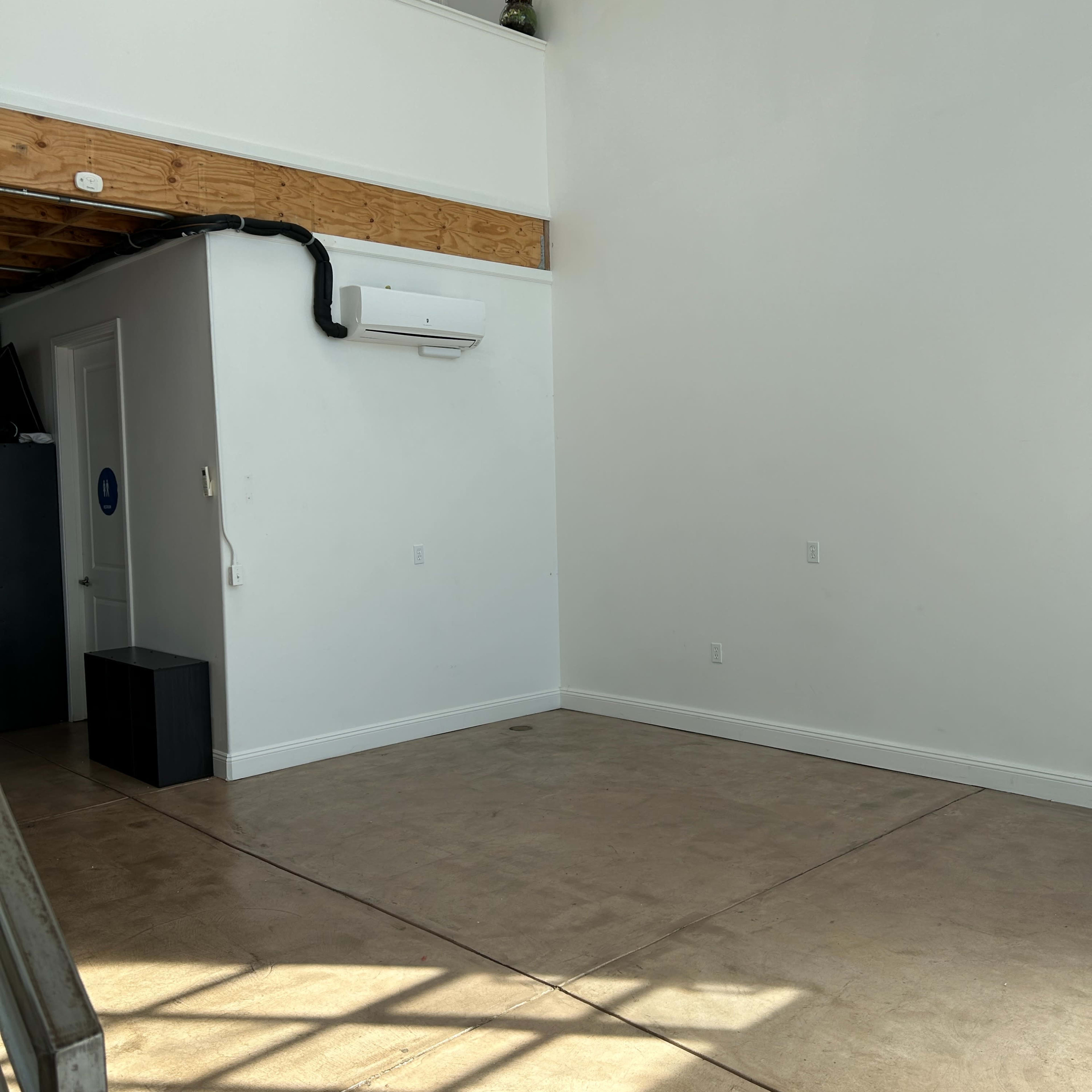 A spacious, empty room with a high ceiling and a concrete floor, illuminated by natural light.