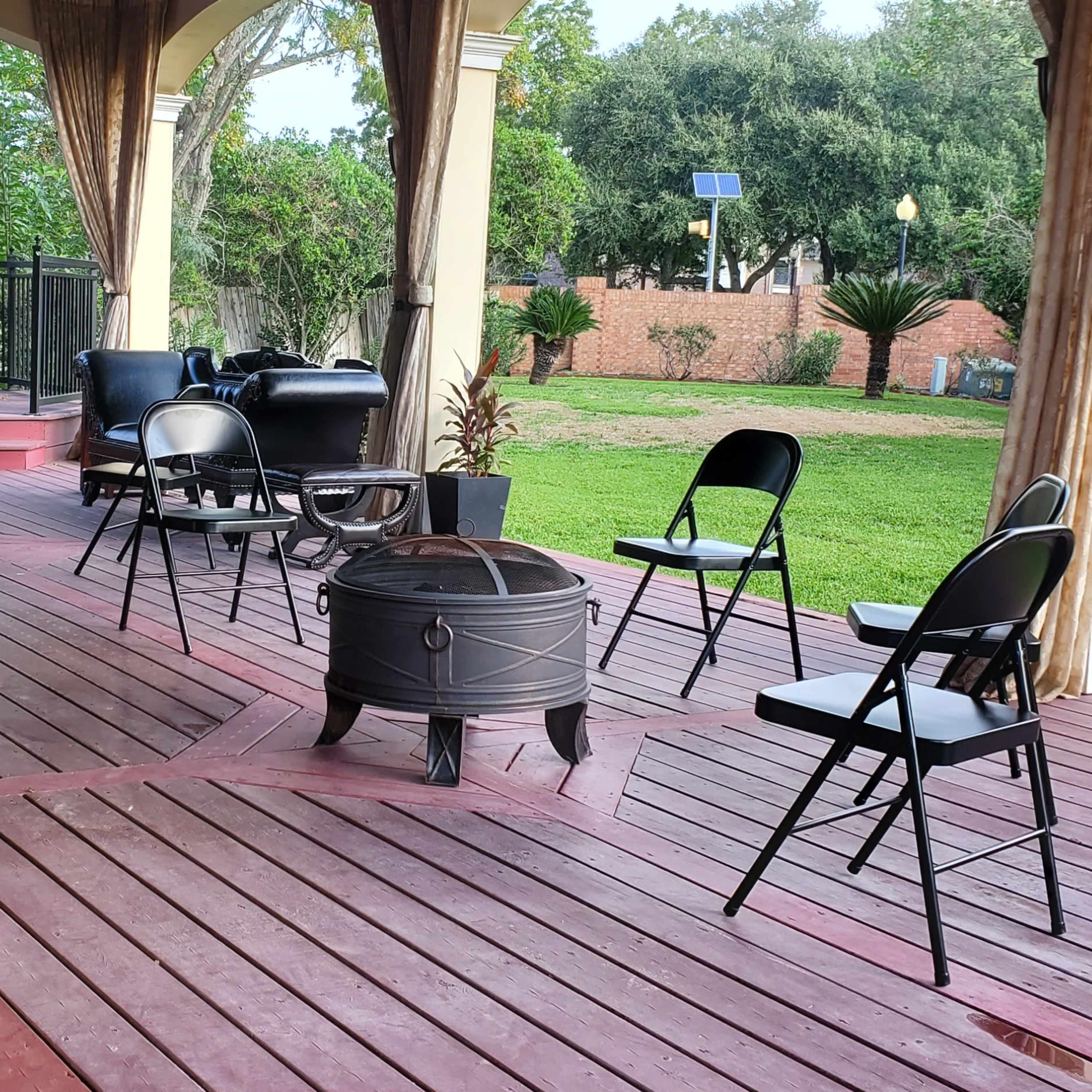 A covered wooden patio with several black folding chairs arranged around a fire pit and potted plants in the background.