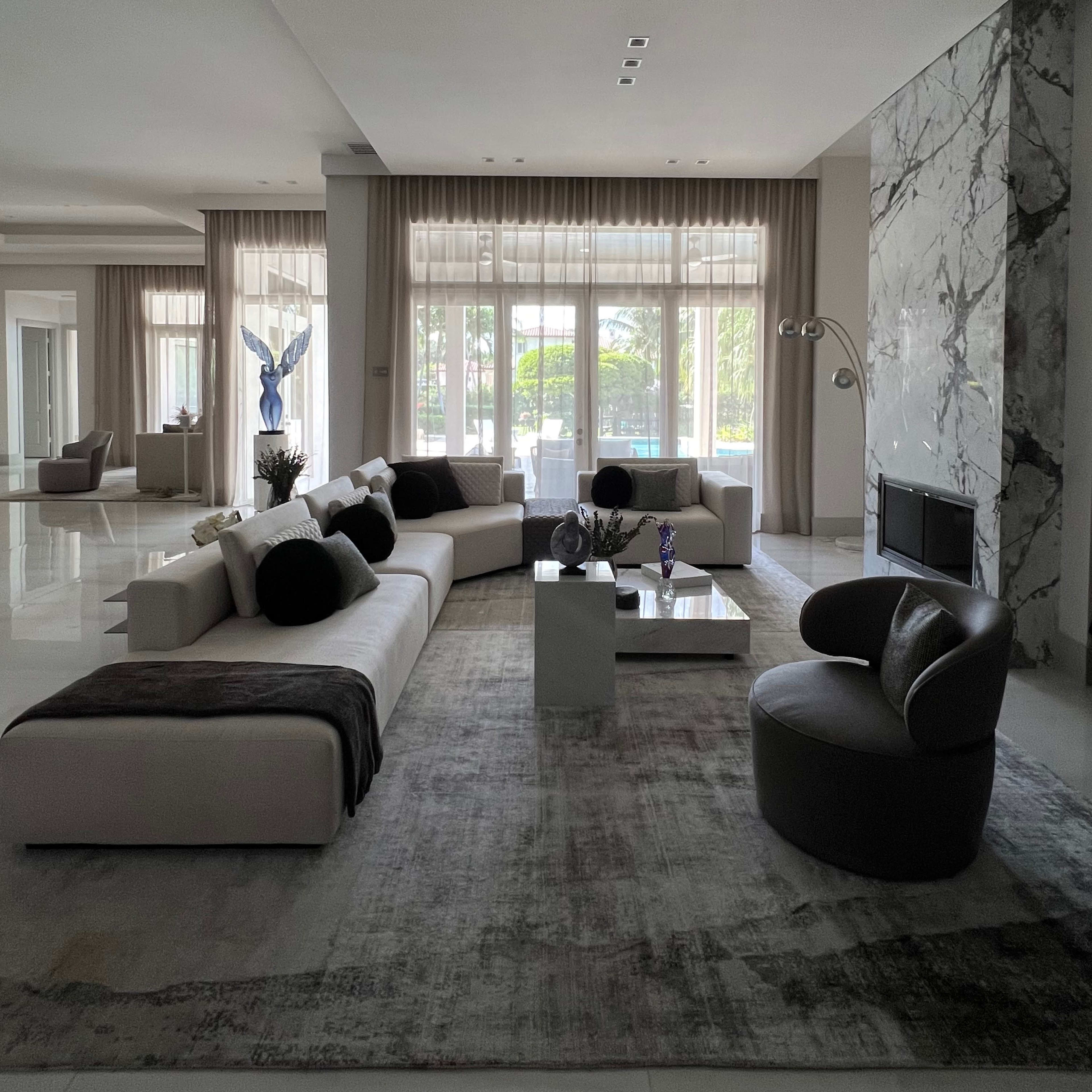 A spacious, modern living room features a beige sectional sofa, a round chair, and a coffee table on a light-colored rug, with large windows overlooking a patio.