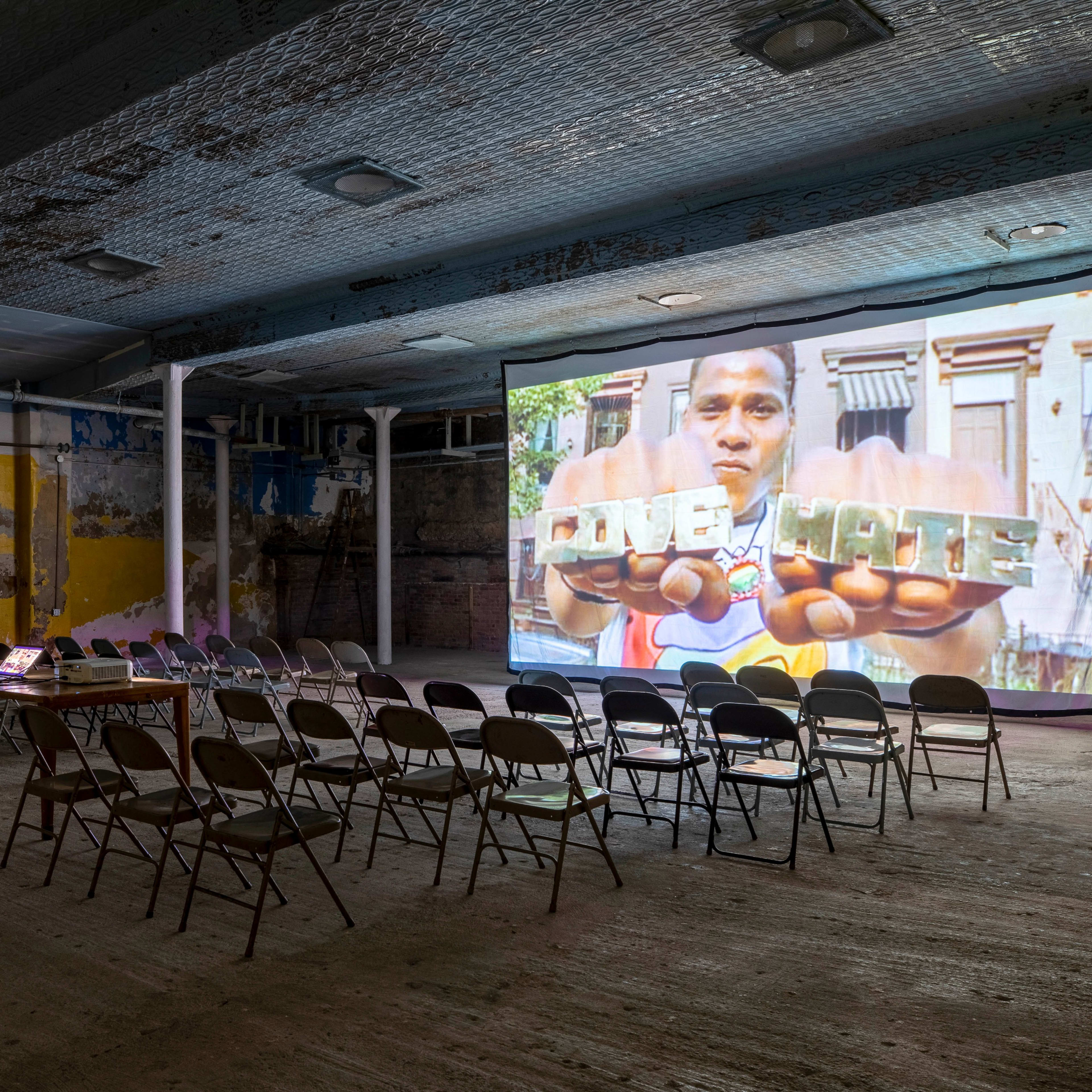 A large projection screen displaying a man holding a sign is set up in an empty room with rows of chairs and exposed brick walls.