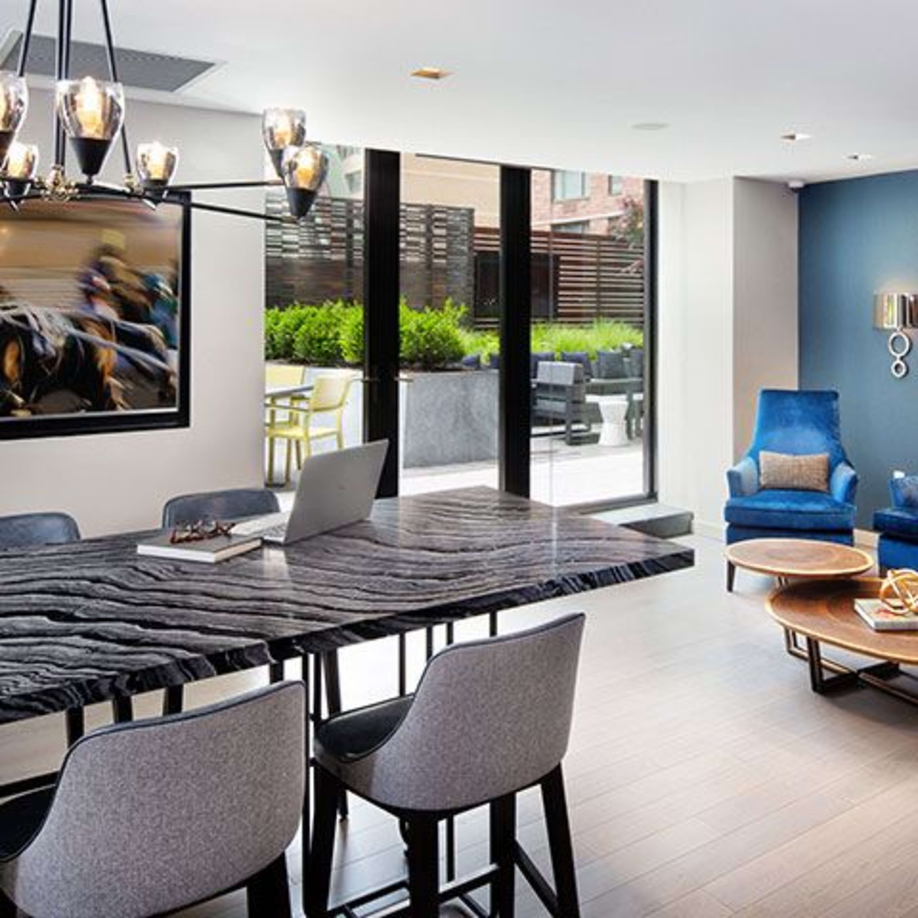 A modern living space features a large dining table, four chairs, and two blue armchairs arranged around a coffee table, with artwork and large windows overlooking a terrace.