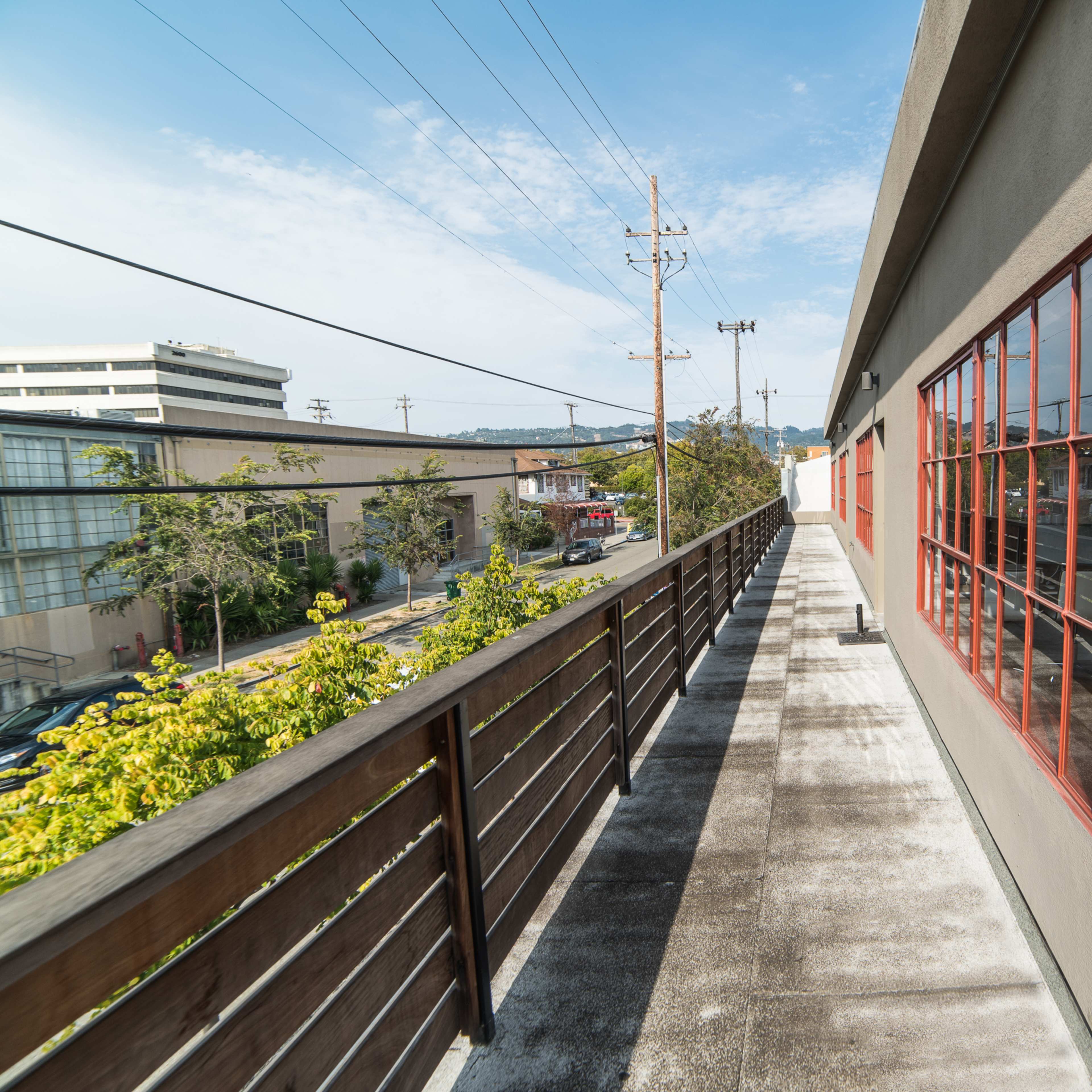 A narrow walkway with a wooden railing runs alongside a building, offering a view of neighboring structures and power lines against a clear sky.