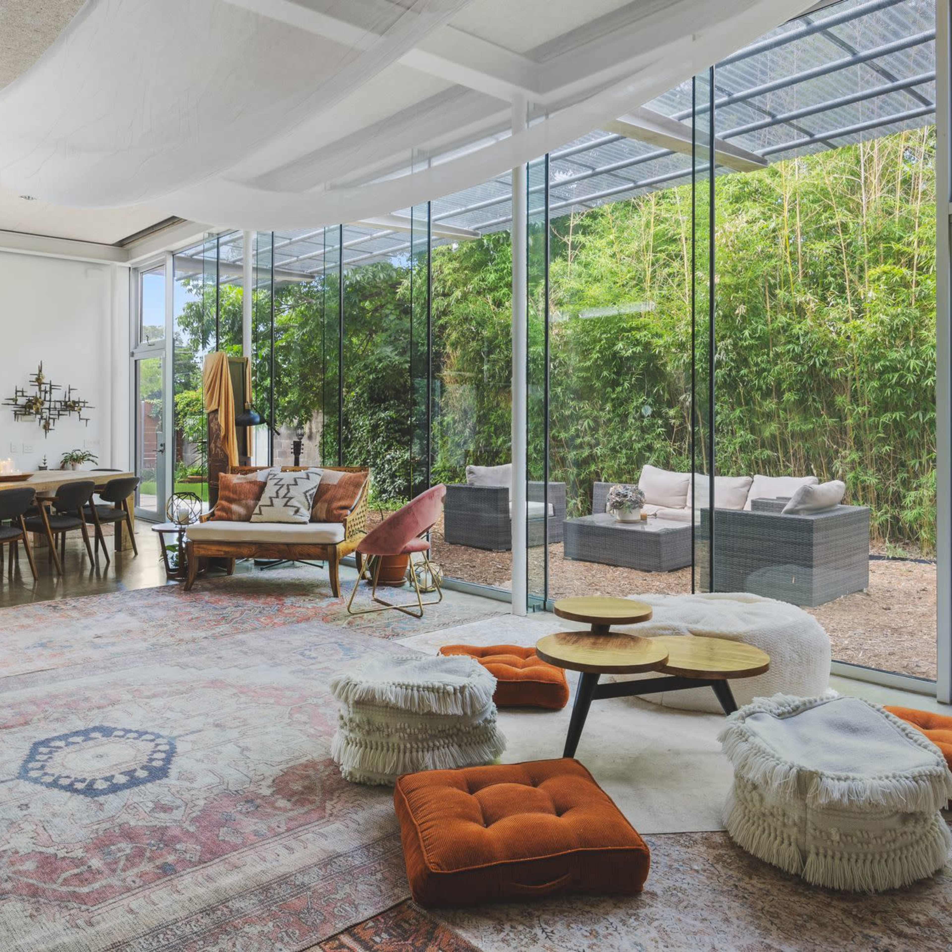 A modern living space features large glass walls with views of a garden, a dining table, comfortable seating, and an area rug.