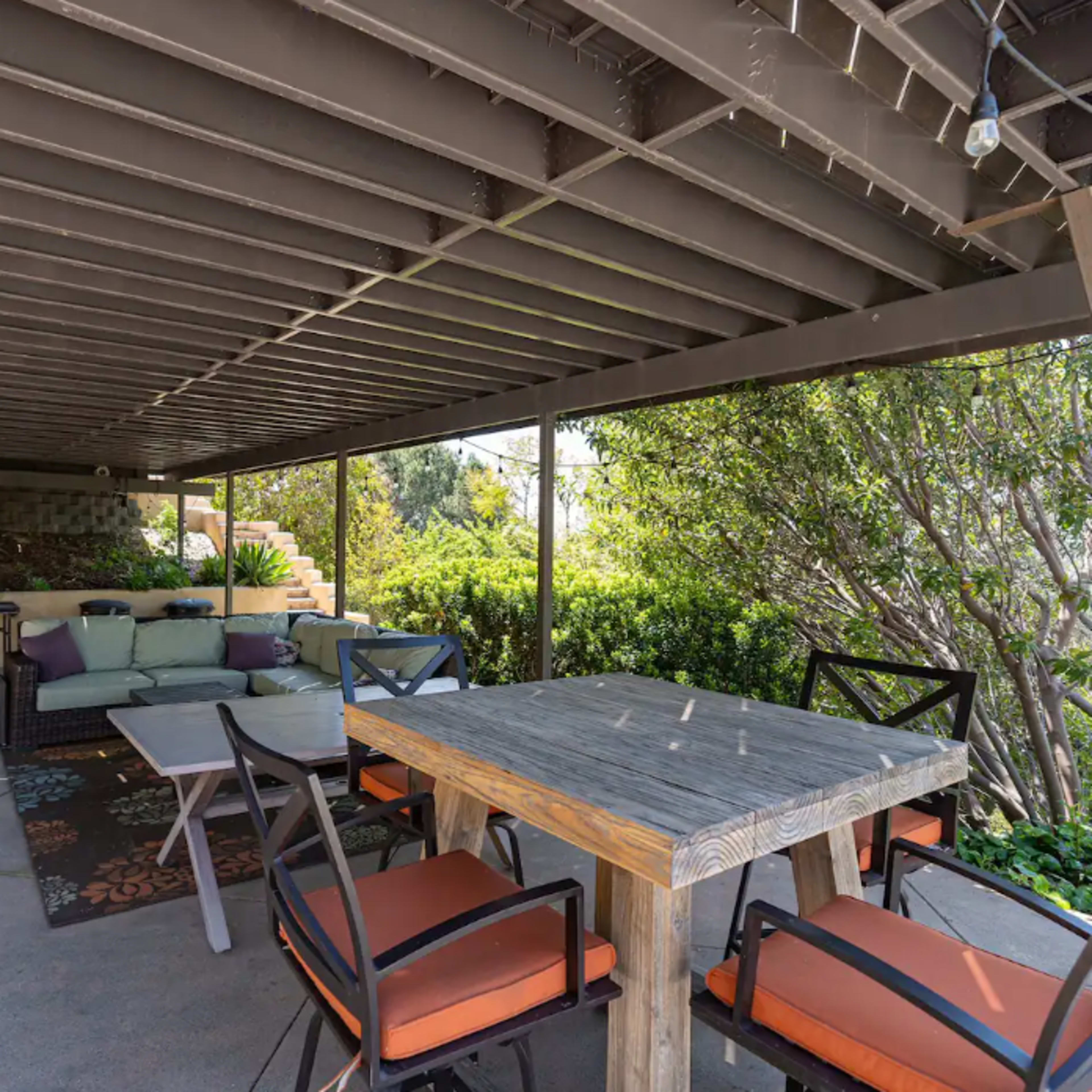 A shaded outdoor patio area with a wooden dining table surrounded by chairs and a lounge section with a sofa and pillows.