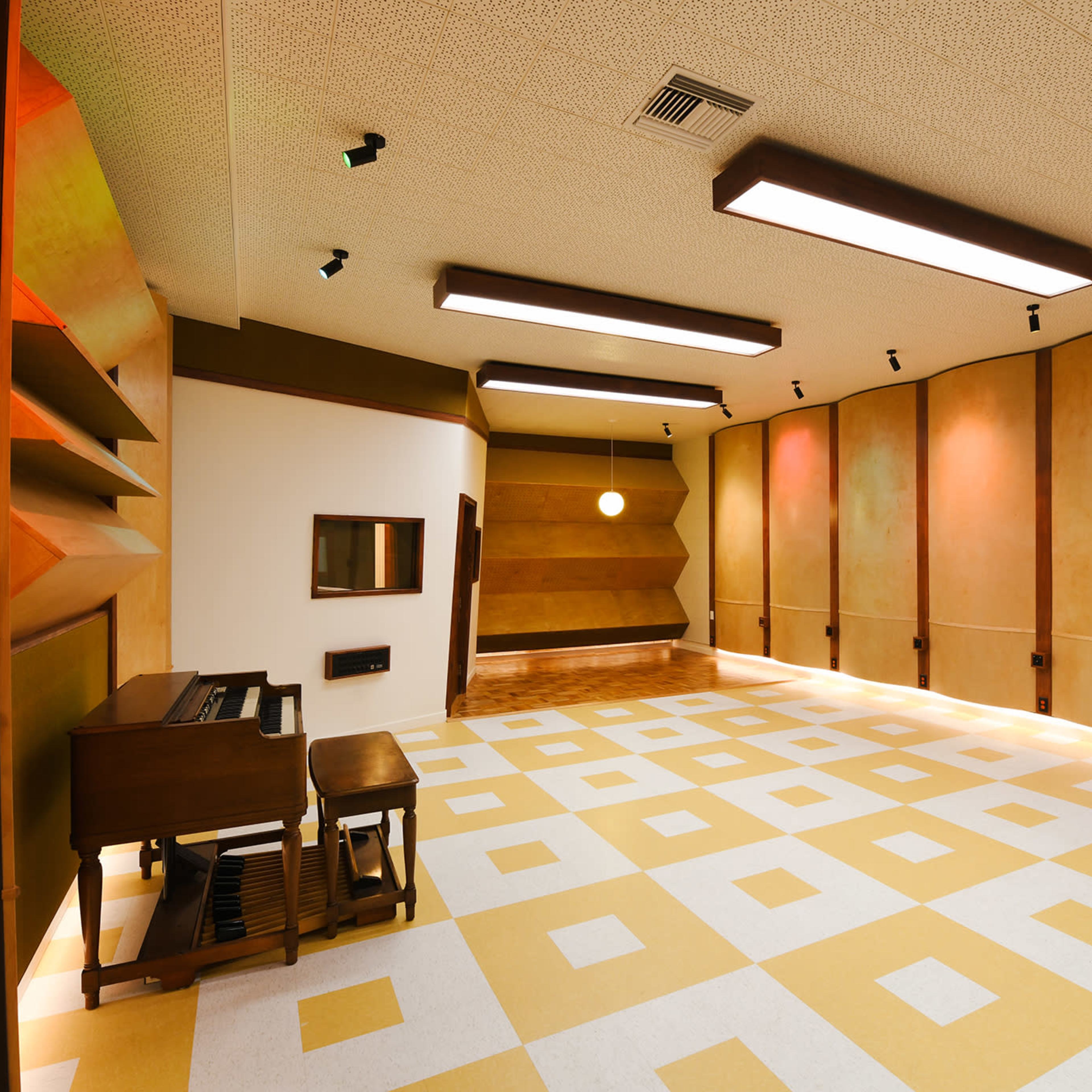 A spacious, well-lit room with a patterned yellow and white floor, featuring two upright pianos and a wood-paneled wall.