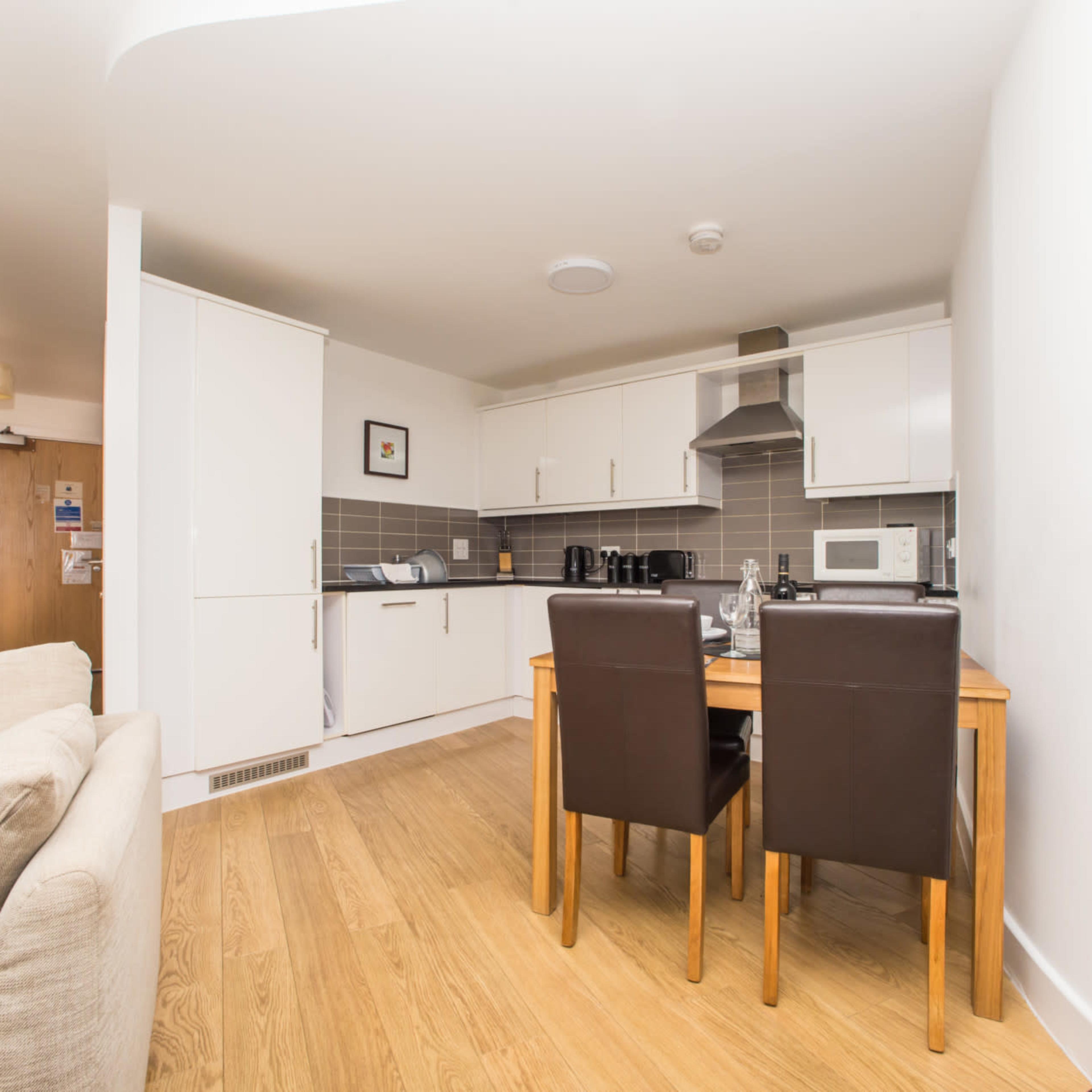 A modern kitchen and dining area with a wooden table and two brown chairs, adjacent to light-colored walls and cabinetry.