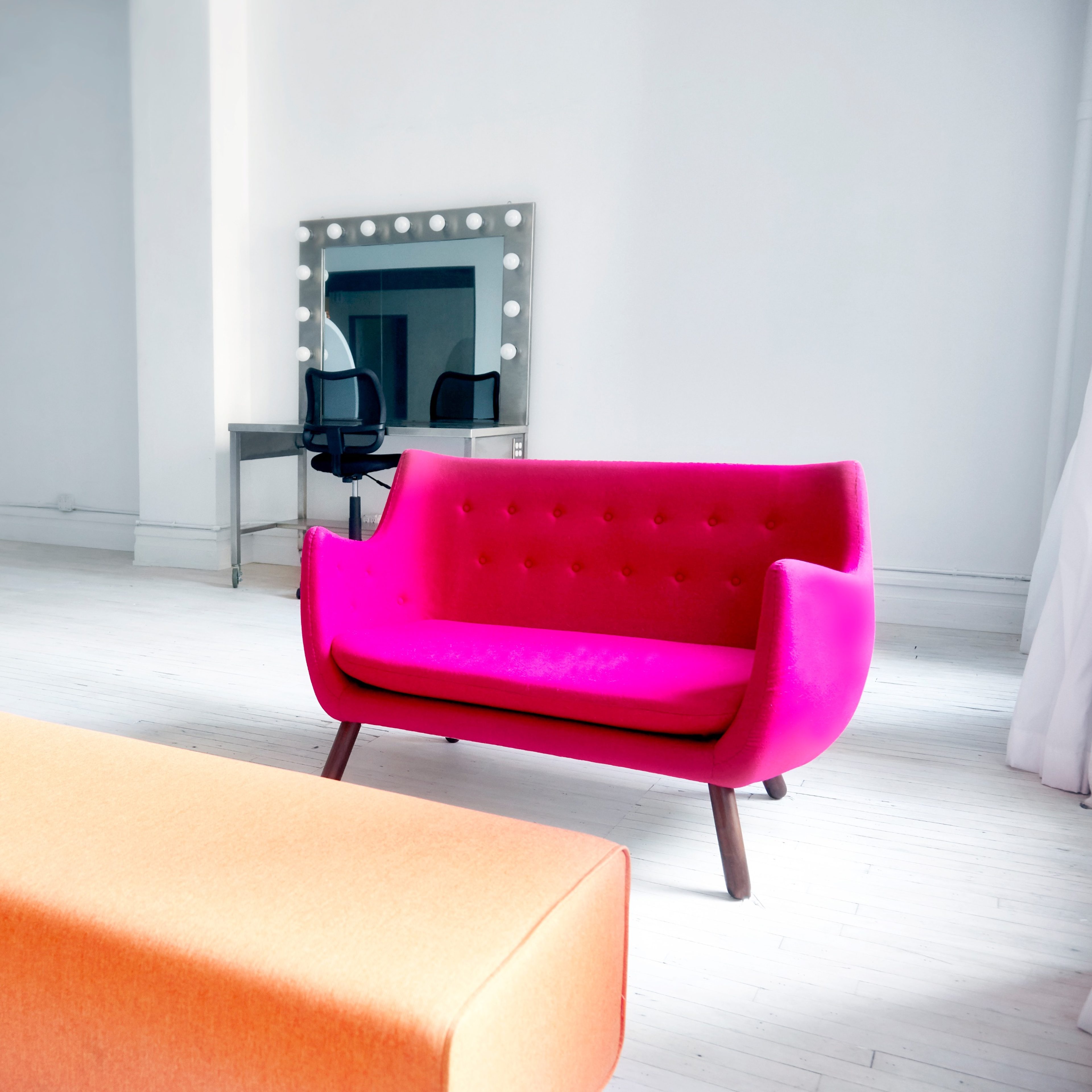 A bright pink sofa and an orange ottoman are positioned in a spacious, minimally decorated room with white walls and wooden floors.