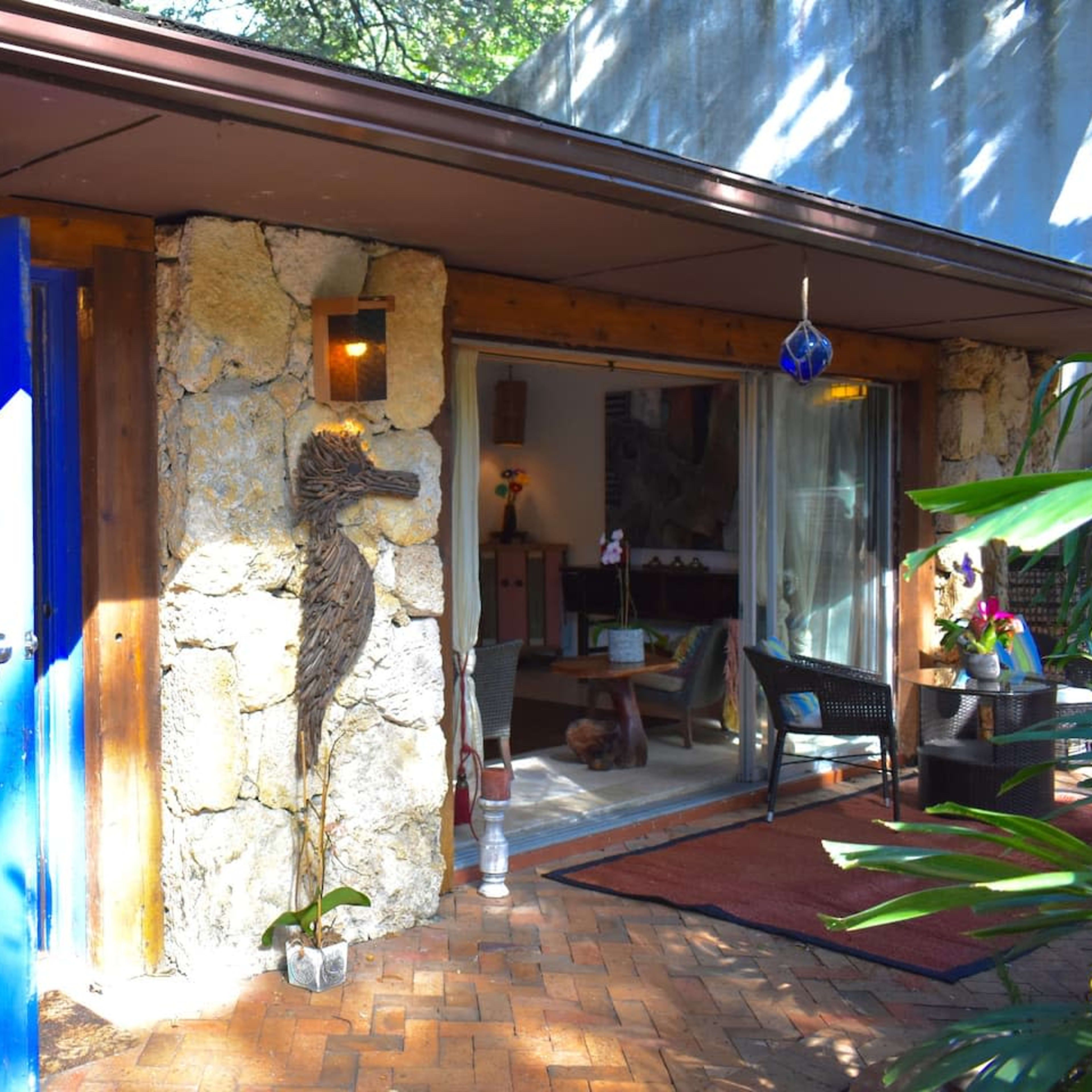 A stone-walled house with a blue door surrounded by greenery and a glimpse of indoor furnishings visible through an open entrance.