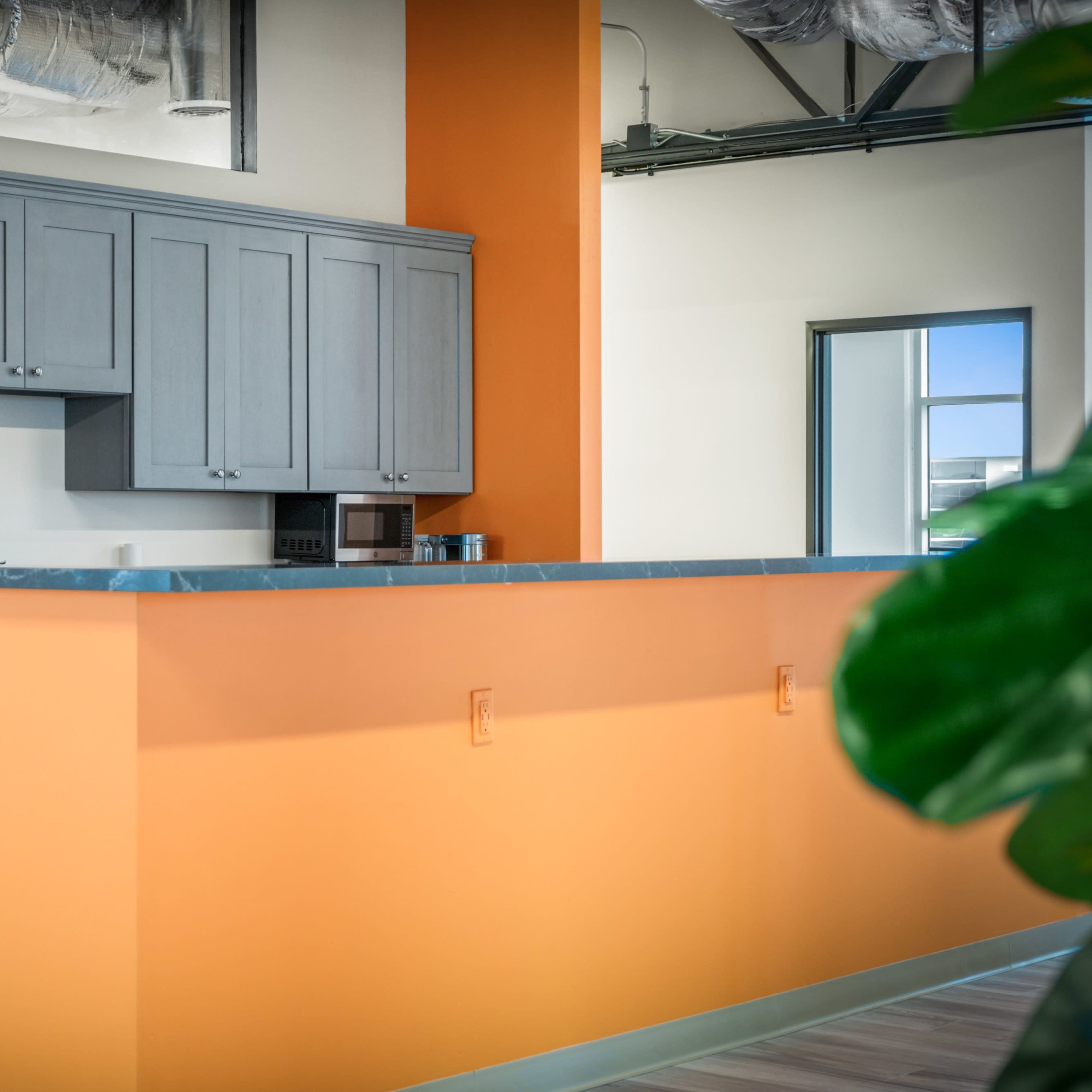 A modern kitchen area with gray cabinets, a stainless steel refrigerator, and an orange wall behind a countertop.