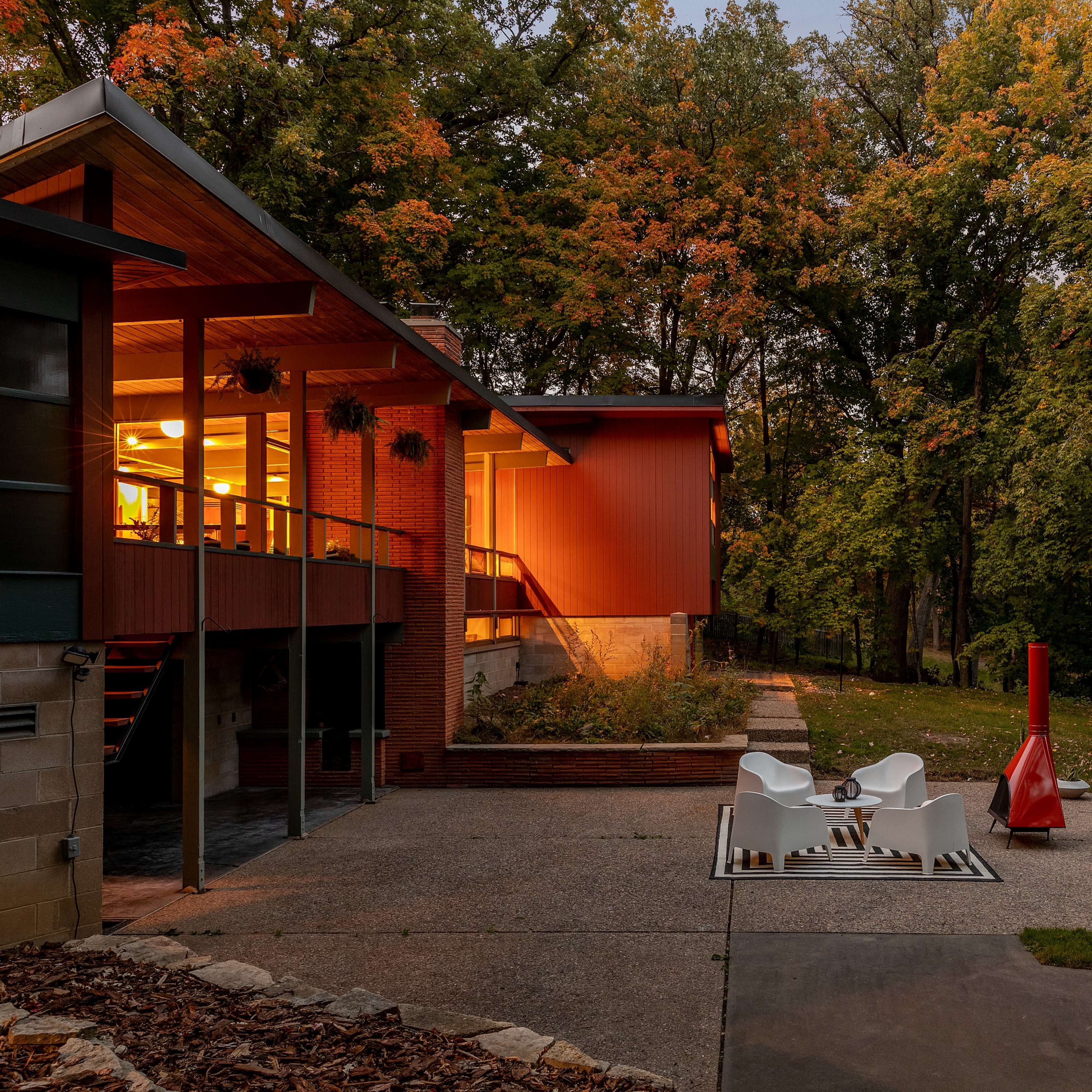 A modern home with a vivid red exterior sits amidst autumn trees, featuring a patio with white chairs and a red outdoor fireplace.